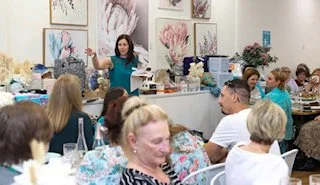 Group of people sitting at tables in a classroom or workshop setting, with a woman standing and speaking at the front, surrounded by art supplies and artwork on the walls.