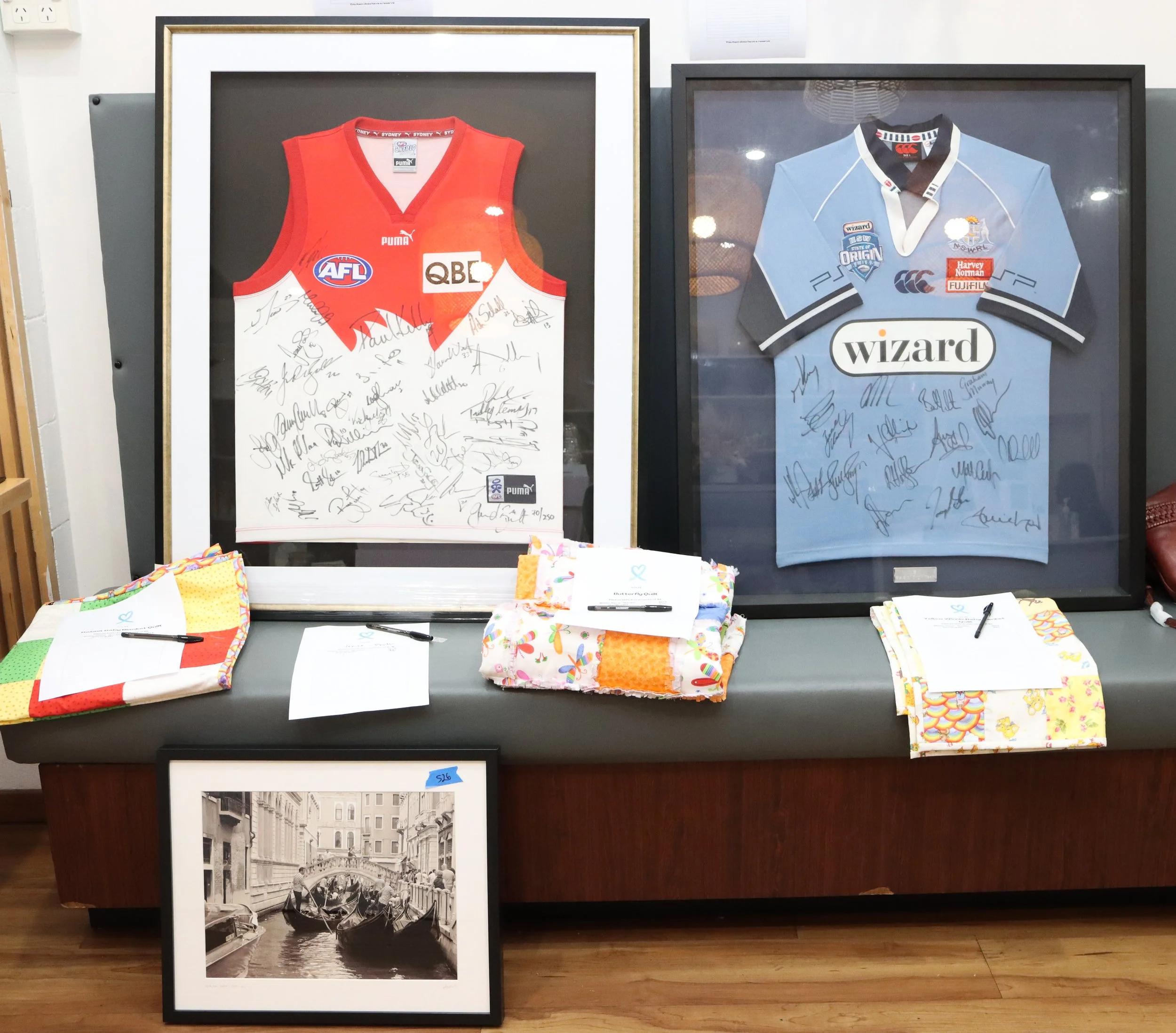 Two framed sports jerseys, one red with AFL and QBE logos, and one blue with Wizard logo, both signed. They are displayed on a grey and black bench with folded fabrics and notes, with a framed black-and-white photo of gondolas in Venice in front.