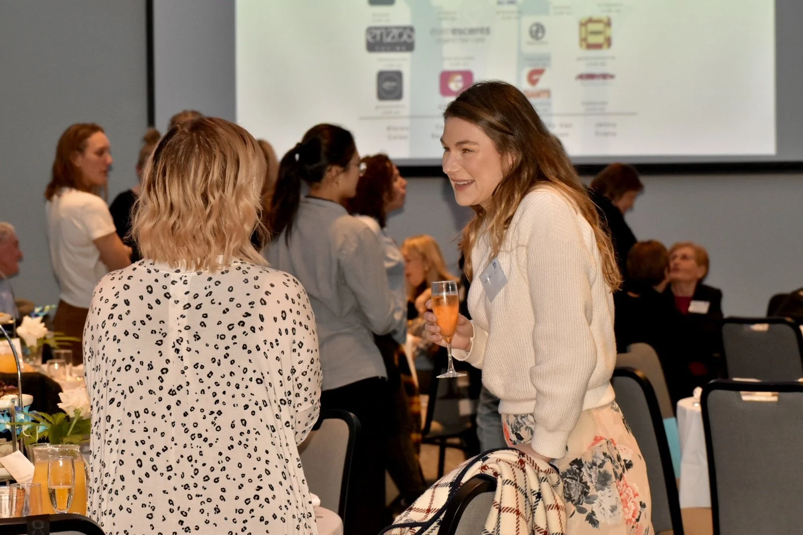 Women chatting at a professional networking event, some holding glasses of drinks, with tables set with flowers and beverages, and a large presentation screen in the background.