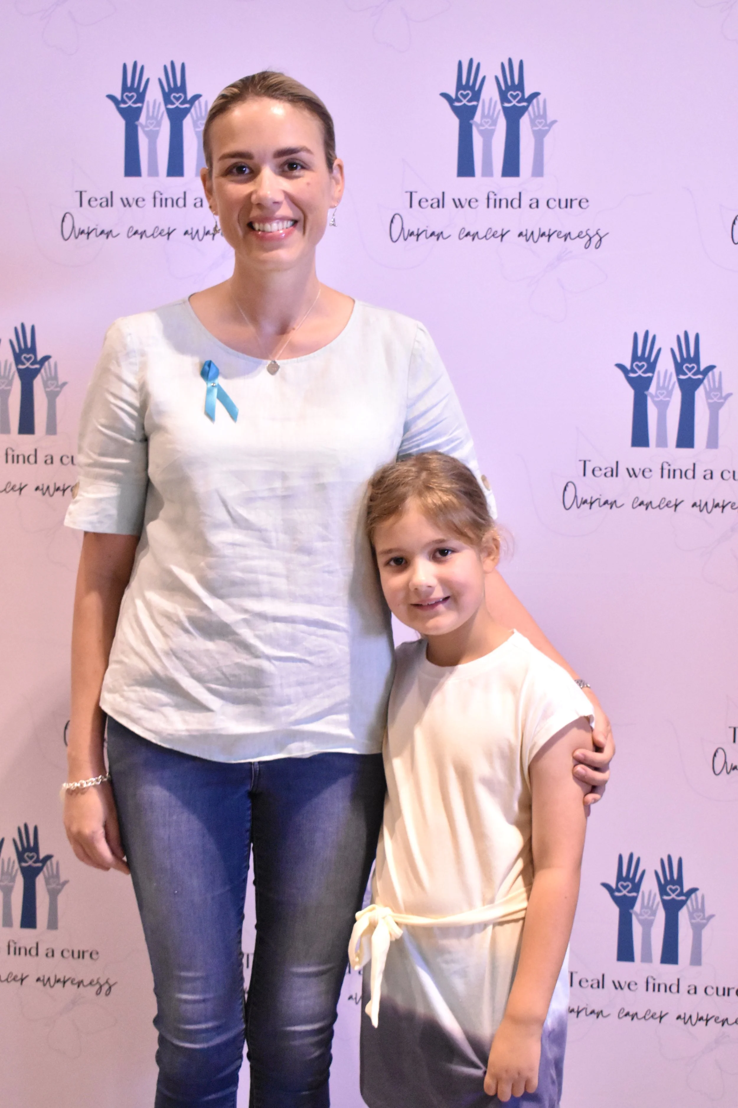 A woman and a young girl standing together in front of a backdrop with breast cancer awareness symbols and text. The woman is wearing a light-colored top with a blue ribbon pin, and the girl is wearing a light-colored dress. Both are smiling.