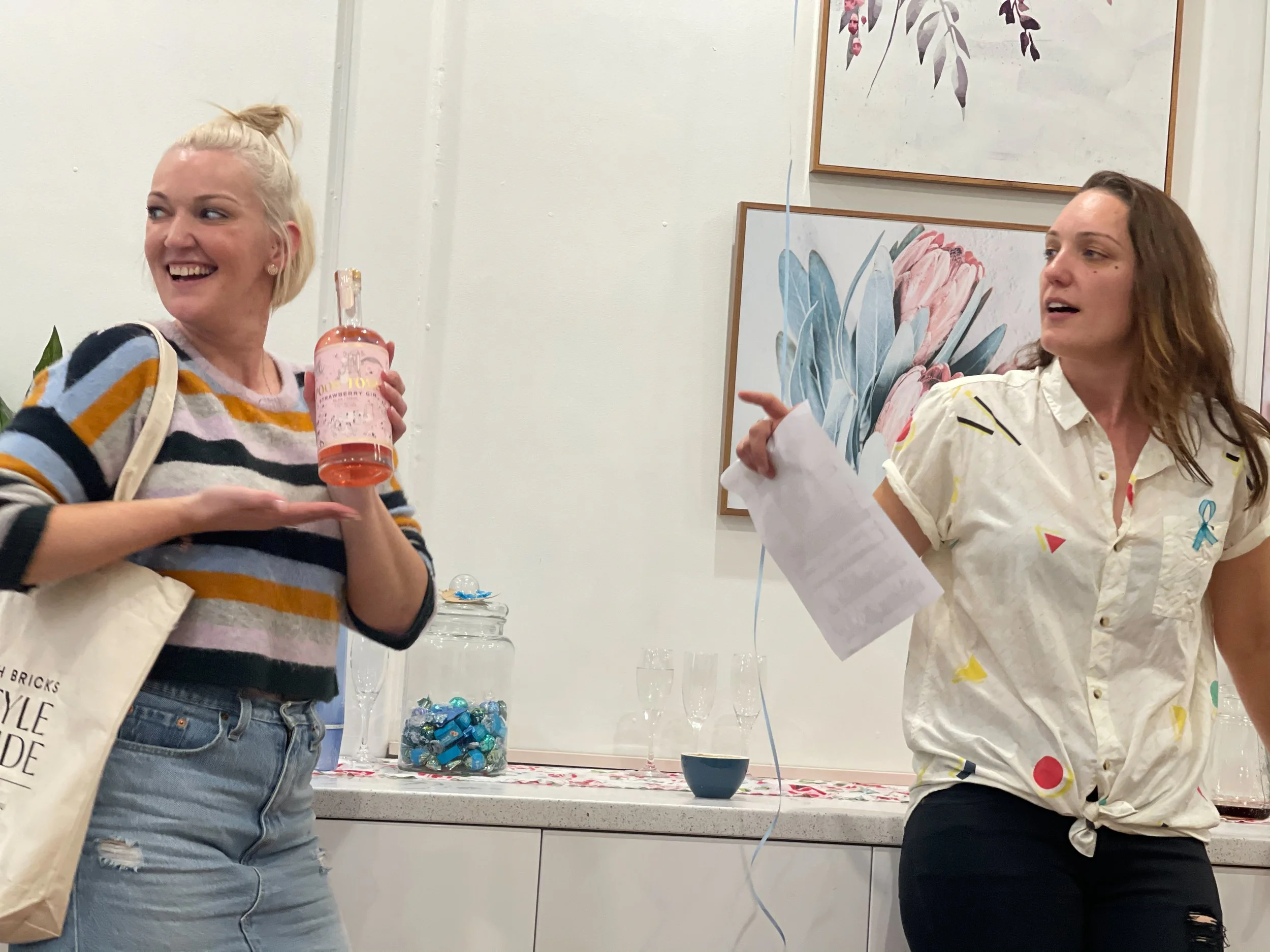 Two women standing in a room, one holding a bottle of pink drink or wine, smiling, and the other pointing and holding a piece of paper, with artwork on the wall behind them.