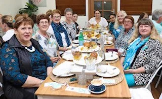 Group of women sitting around a dining table with a tiered tray of food and drinks, indoors at a social gathering or celebration.