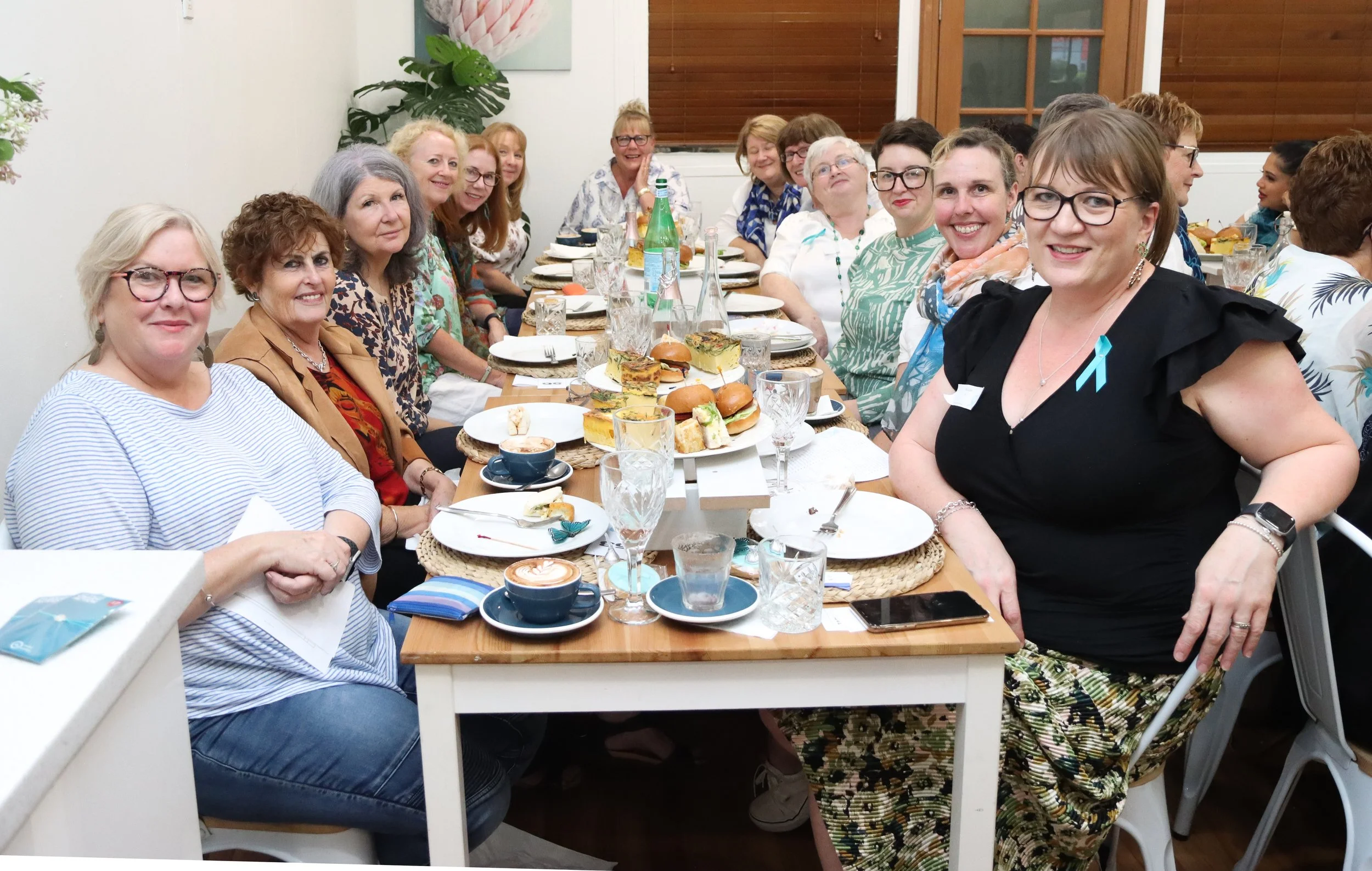 Group of women sitting around a table at a social gathering, with food and drinks on the table, smiling and looking at the camera.