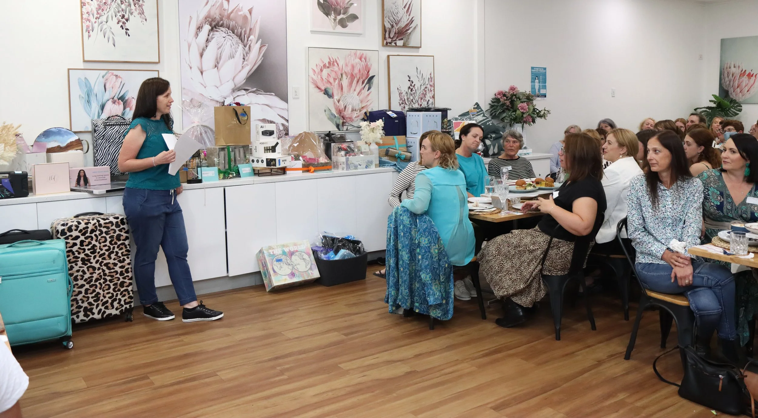A woman with long brown hair stands at the front of a room, speaking to a seated audience of women at tables, some eating and drinking. The room has floral artwork on the walls and various gift bags and boxes on a sideboard.