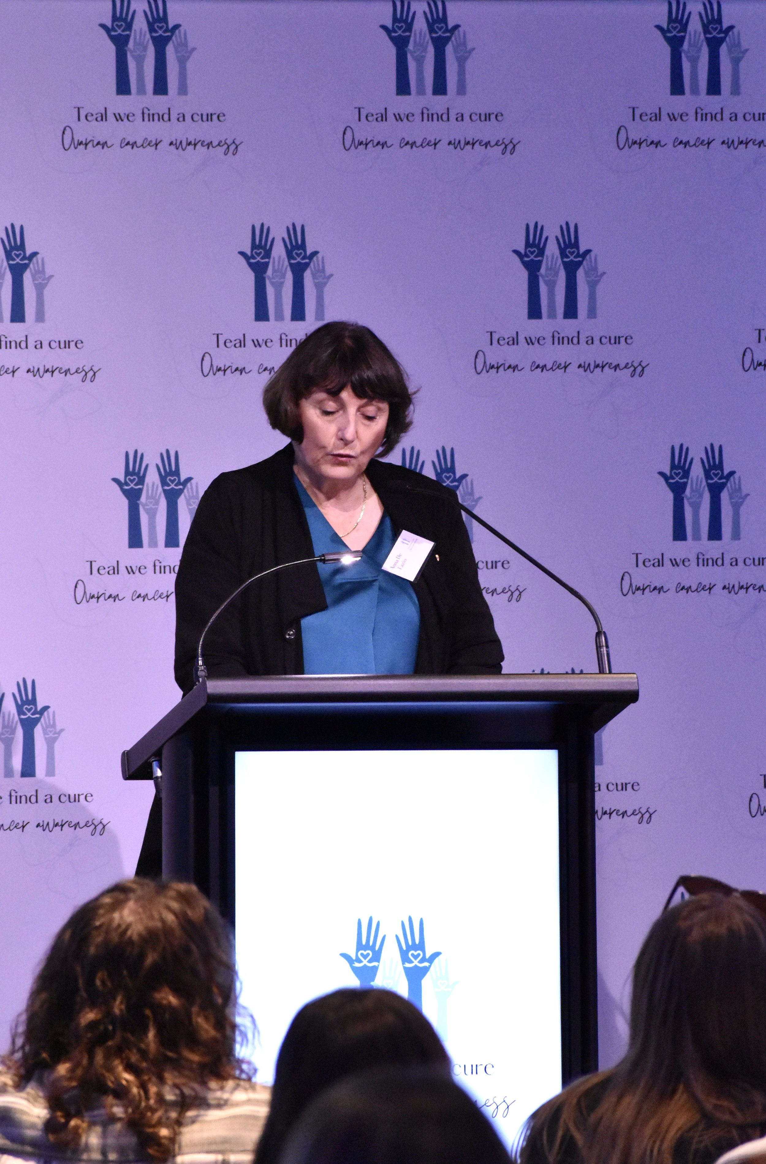 A woman speaking at a podium during an ovarian cancer awareness event, with a backdrop displaying the logo and slogan 'Teal we find a cure, Ovarian cancer awareness.'