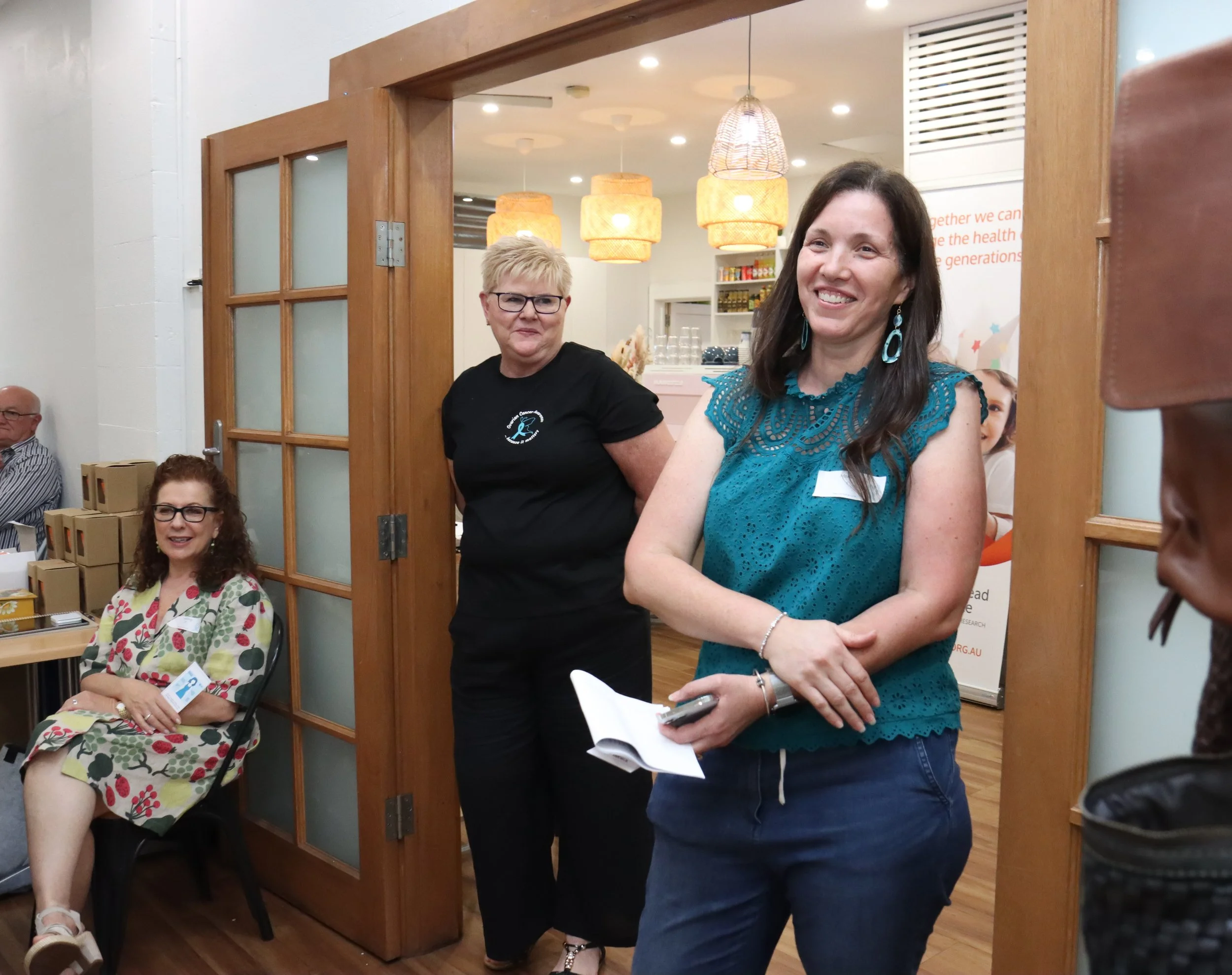 Women smiling and standing in a room with a woman sitting at a table, with wooden doors and hanging lights in the background.