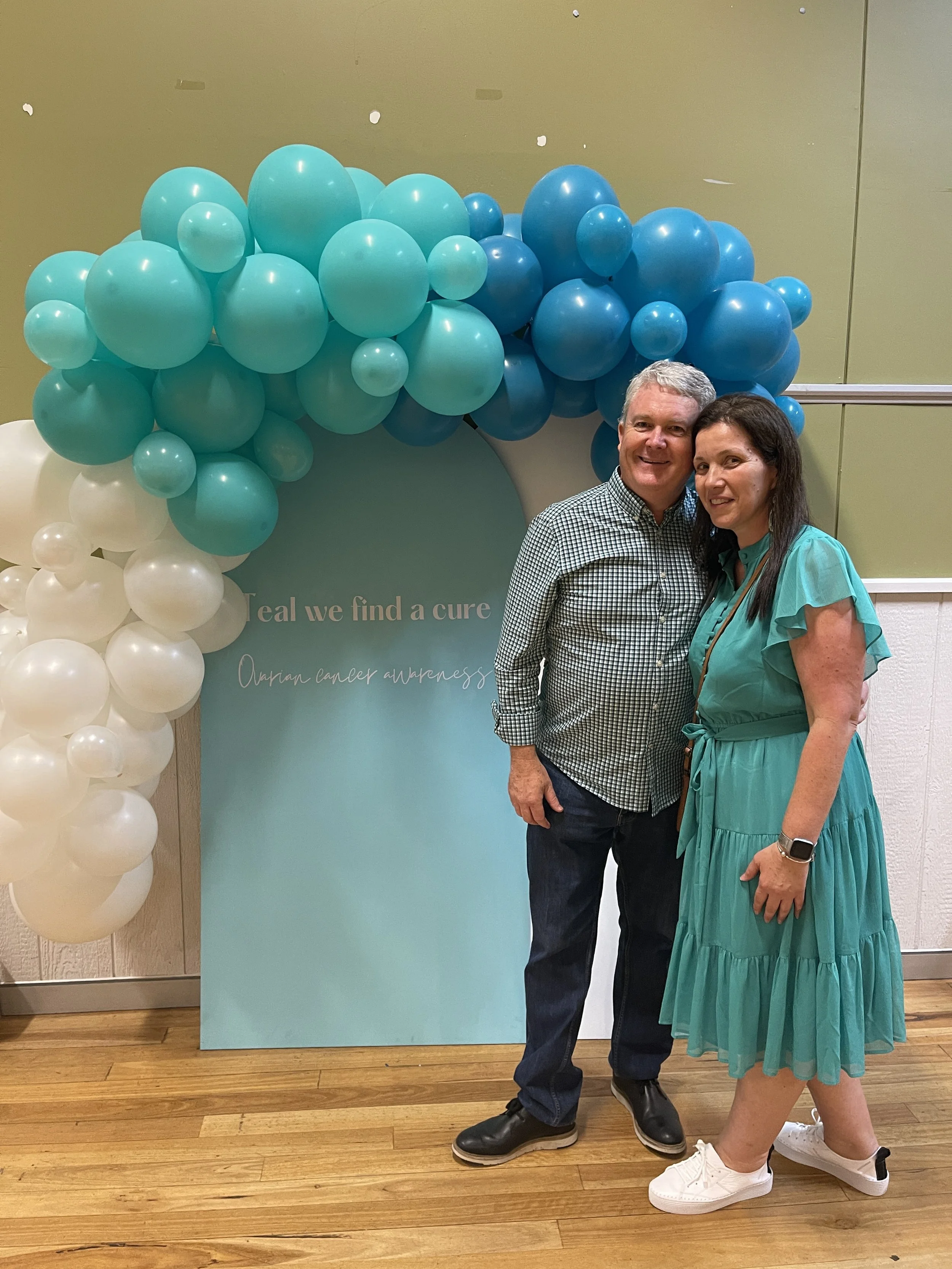A man and woman standing side by side smiling in front of teal-colored backdrop with blue and white balloons arching over it, at a cancer awareness event.