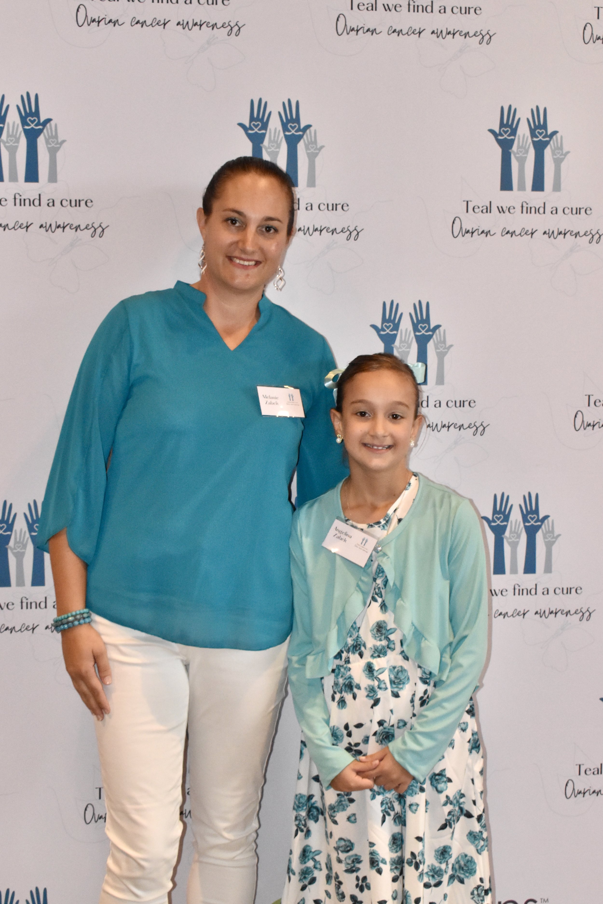 A woman and a girl standing in front of a backdrop with the logo of a hands with hearts and the text 'Teal we find a cure, ovarian cancer awareness.' The woman is wearing a turquoise top and white pants, and the girl is wearing a floral dress with a 