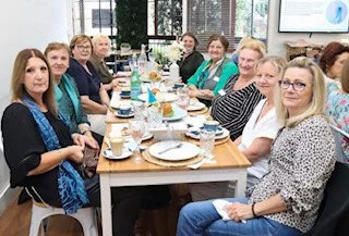 Group of women sitting around a long table during a meal or meeting in a bright room.