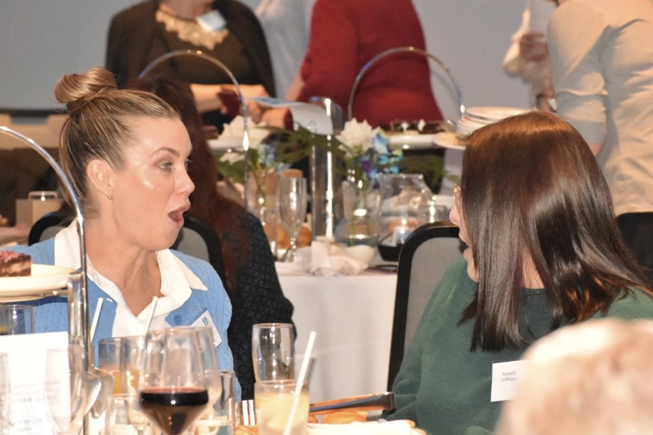 Two women engaged in conversation at a banquet table. The woman on the left has her hair in a bun and is wearing a blue and white jacket, while the woman on the right has straight brown hair and is wearing a dark green top. They are surrounded by foo