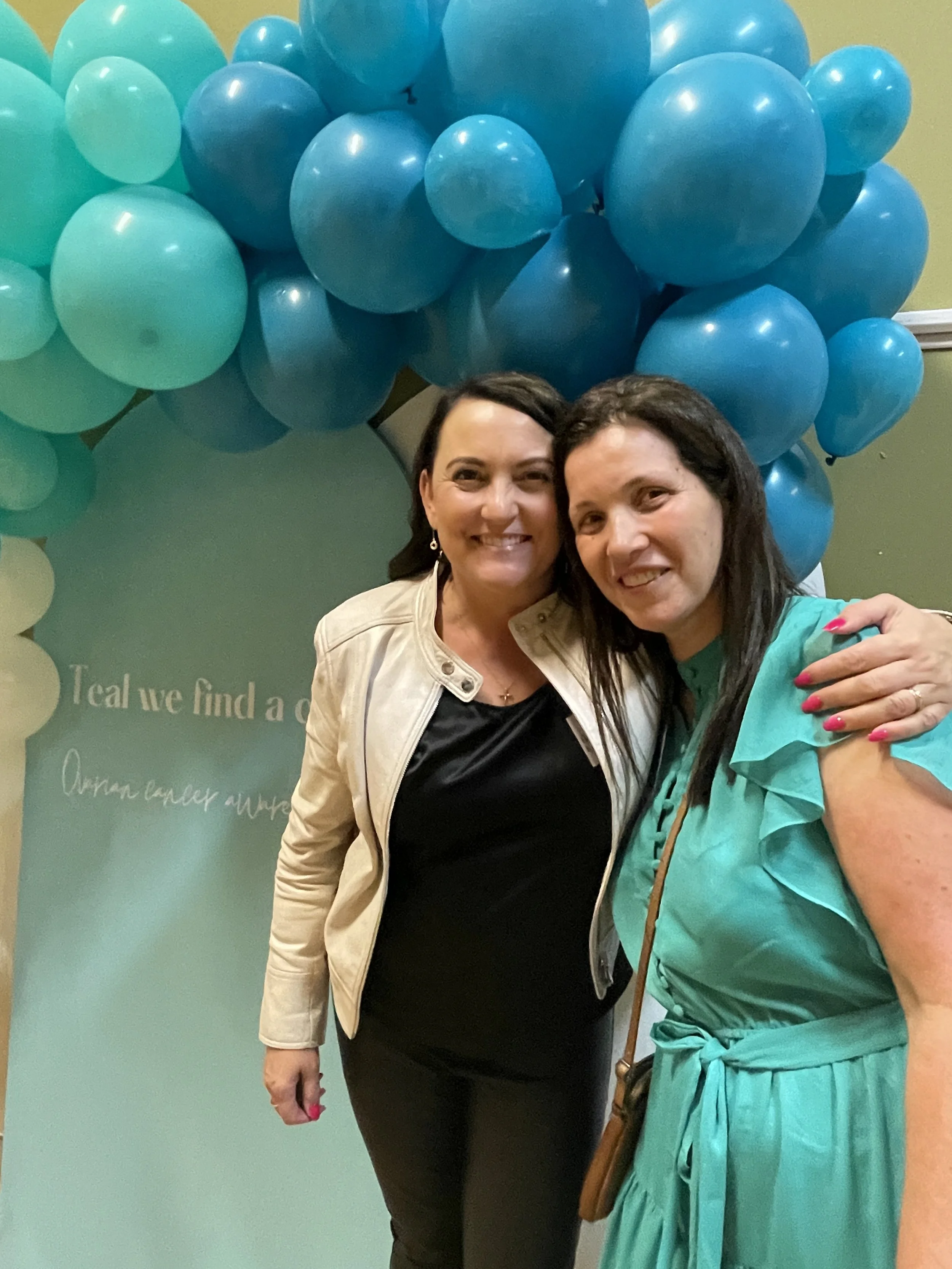 Two women smiling, standing close together with their arms around each other, in front of a balloon arrangement in shades of blue.