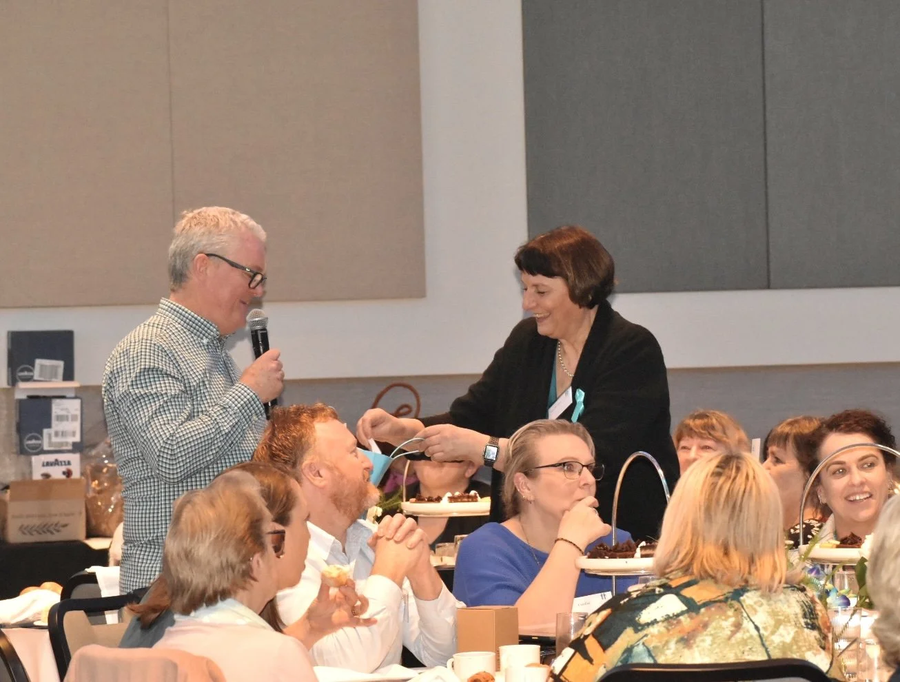 A group of women seated at a banquet table with food and drinks, with a woman in a black jacket and a teal ribbon presenting an award or gift to a man holding a microphone, during a formal event.