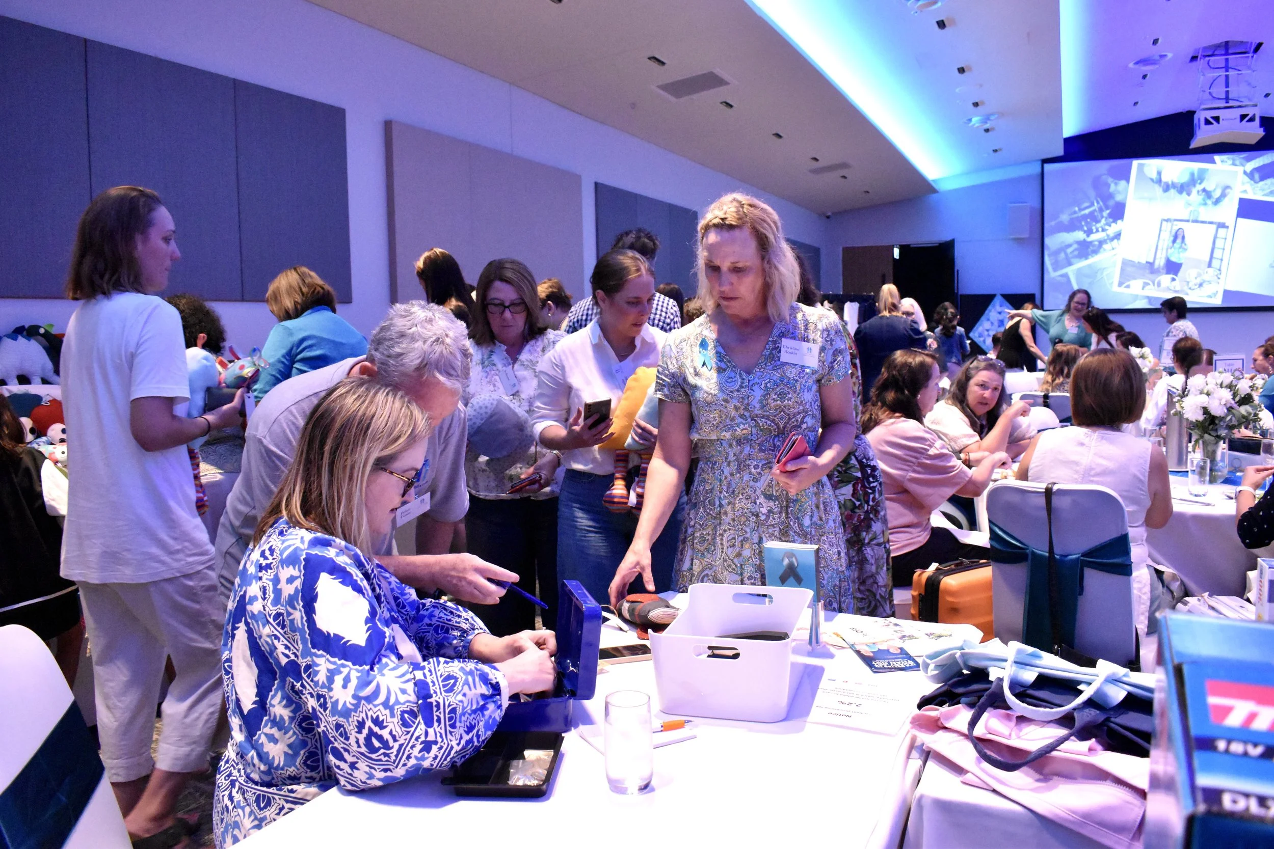 People gathered around a table at a conference or event, engaging in conversation, browsing items, and using devices, with a stage or presentation screen in the background.