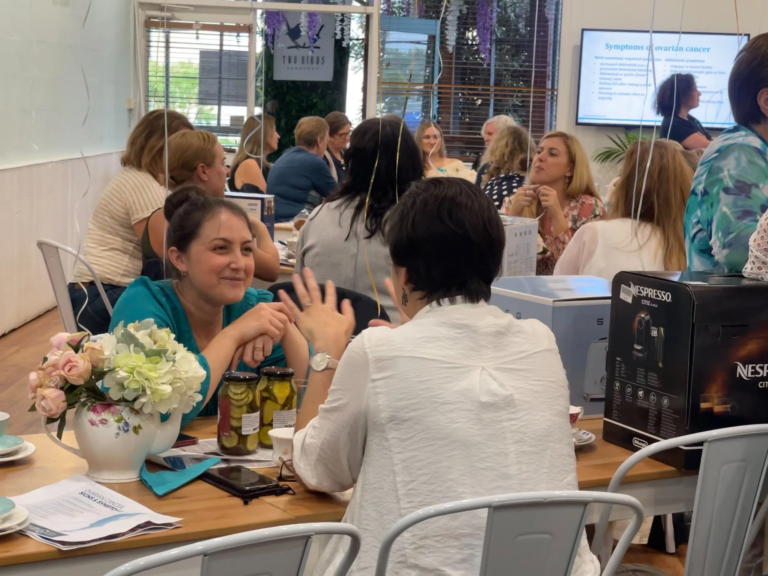 Group of women sitting around a table at a conference or seminar, engaging in discussion, with a presentation on a monitor in the background, and flowers and various items on the table.