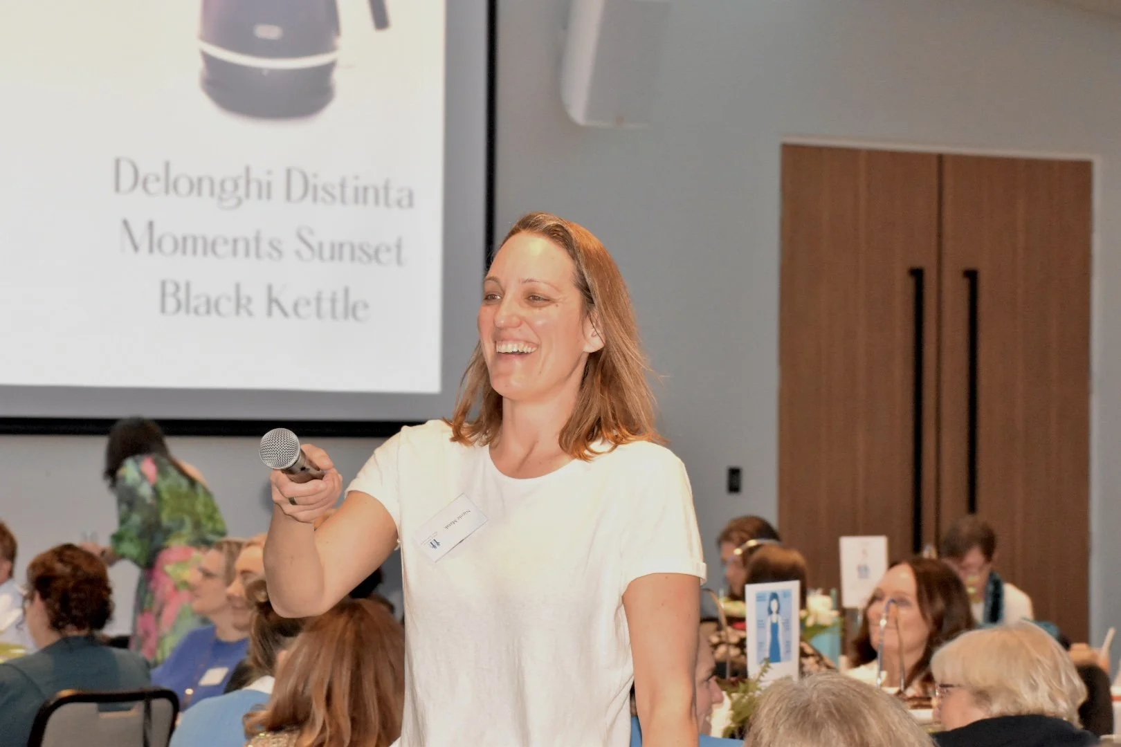 A woman is smiling and holding a microphone at a conference or gathering with many people seated at round tables. A large screen behind her displays the text 'Delonghi Distinta Moments Sunset Black kettle'.