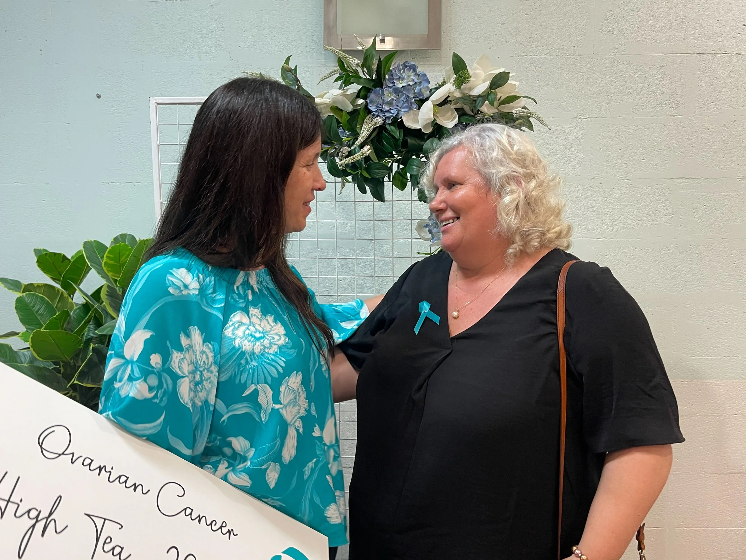 Two women smiling and greeting each other at an ovarian cancer awareness event, standing near a floral arrangement with blue and white flowers.