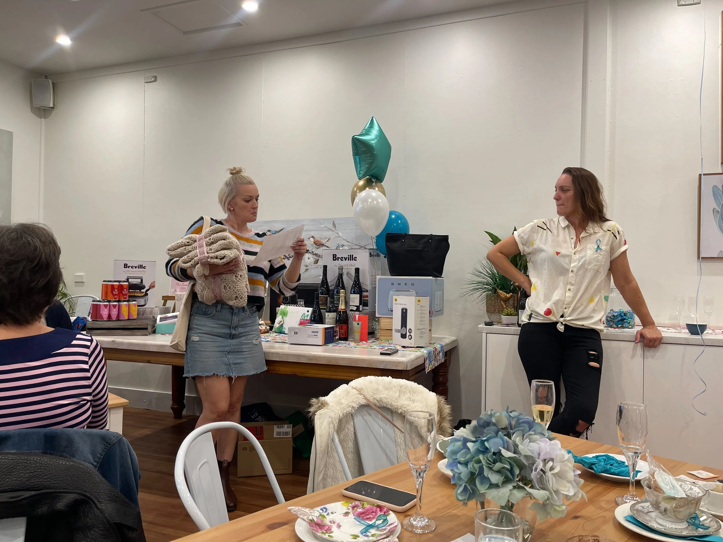 Women are standing in a room decorated with balloons, plants, and streamers. One woman, holding scarves and reading, is on the left, while another woman leans against the wall on the right. A table in front has various gifts and bottles.