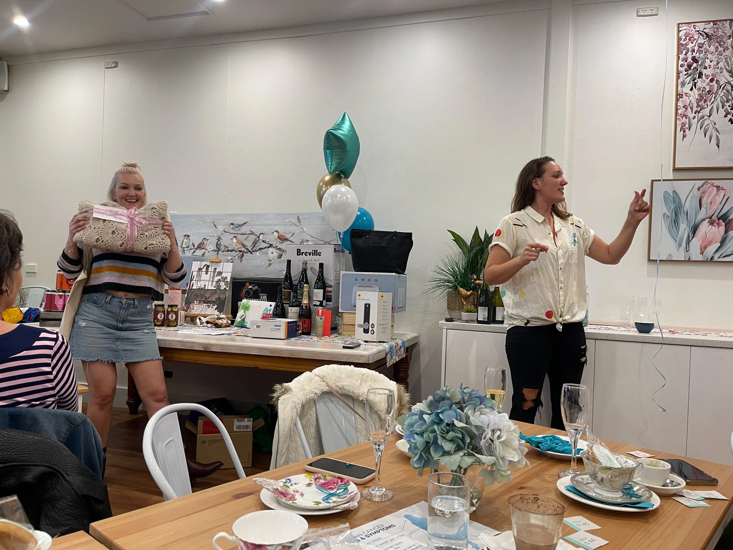 Two women are standing at a party with gifts, balloons, and drinks. One woman is holding a wrapped present, and the other is speaking and gesturing. The table has flowers, glasses, and tableware.