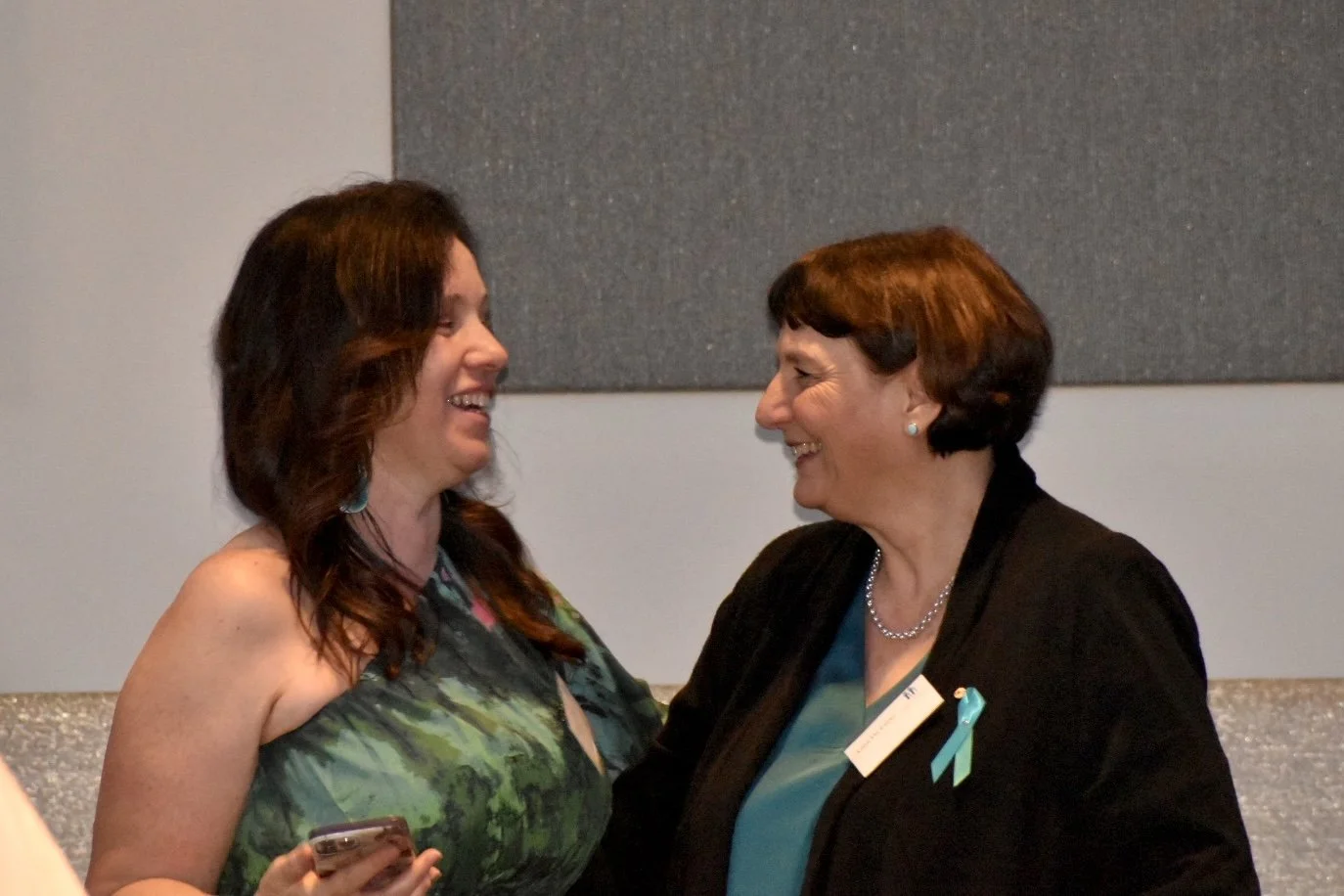 Two women smiling and facing each other at a formal event, with one holding a phone and both wearing name tags and ribbons on their clothing.