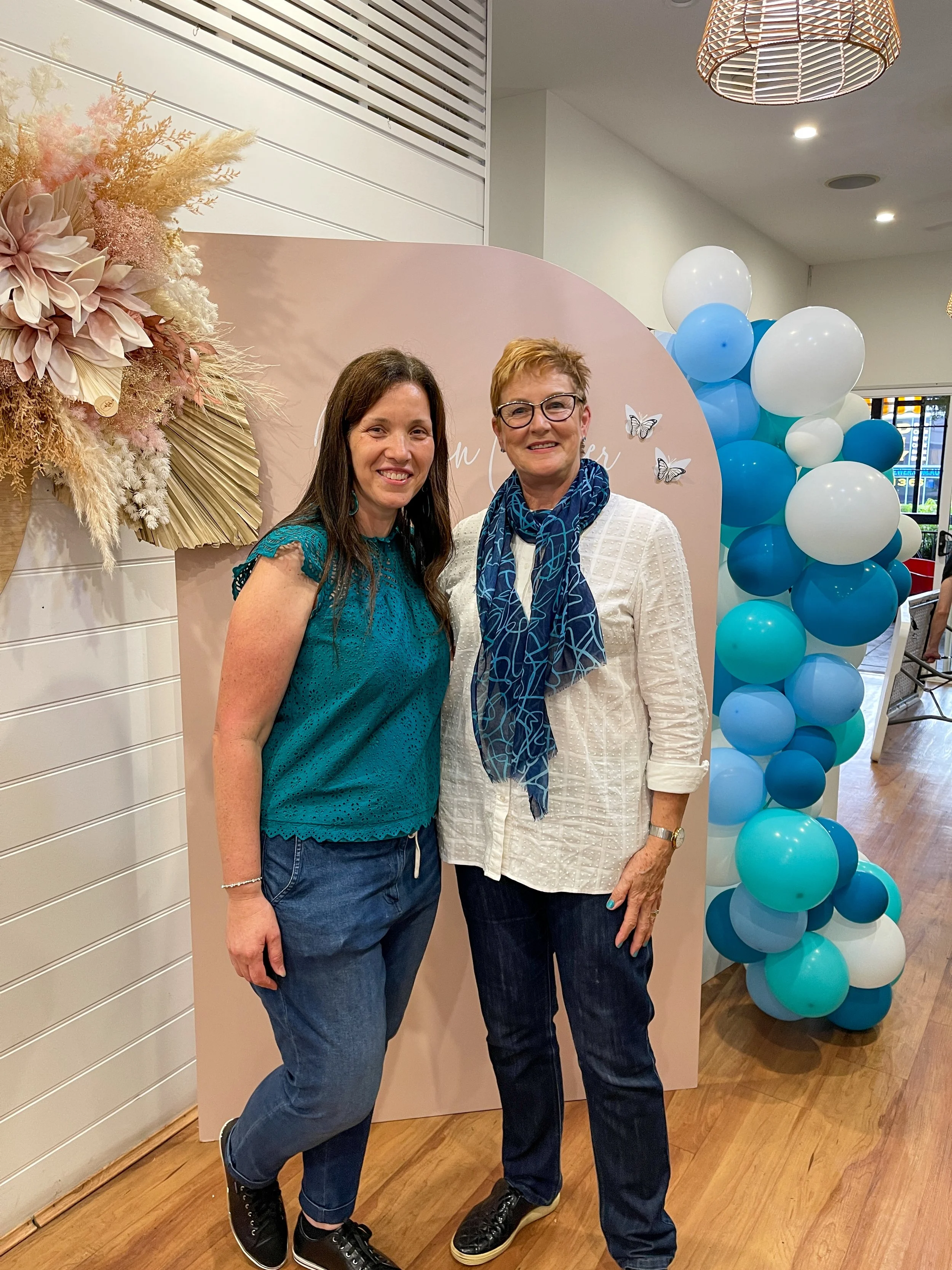 Two women standing together indoors near a pink backdrop decorated with flowers and butterflies, with a cluster of blue and white balloons on the side.