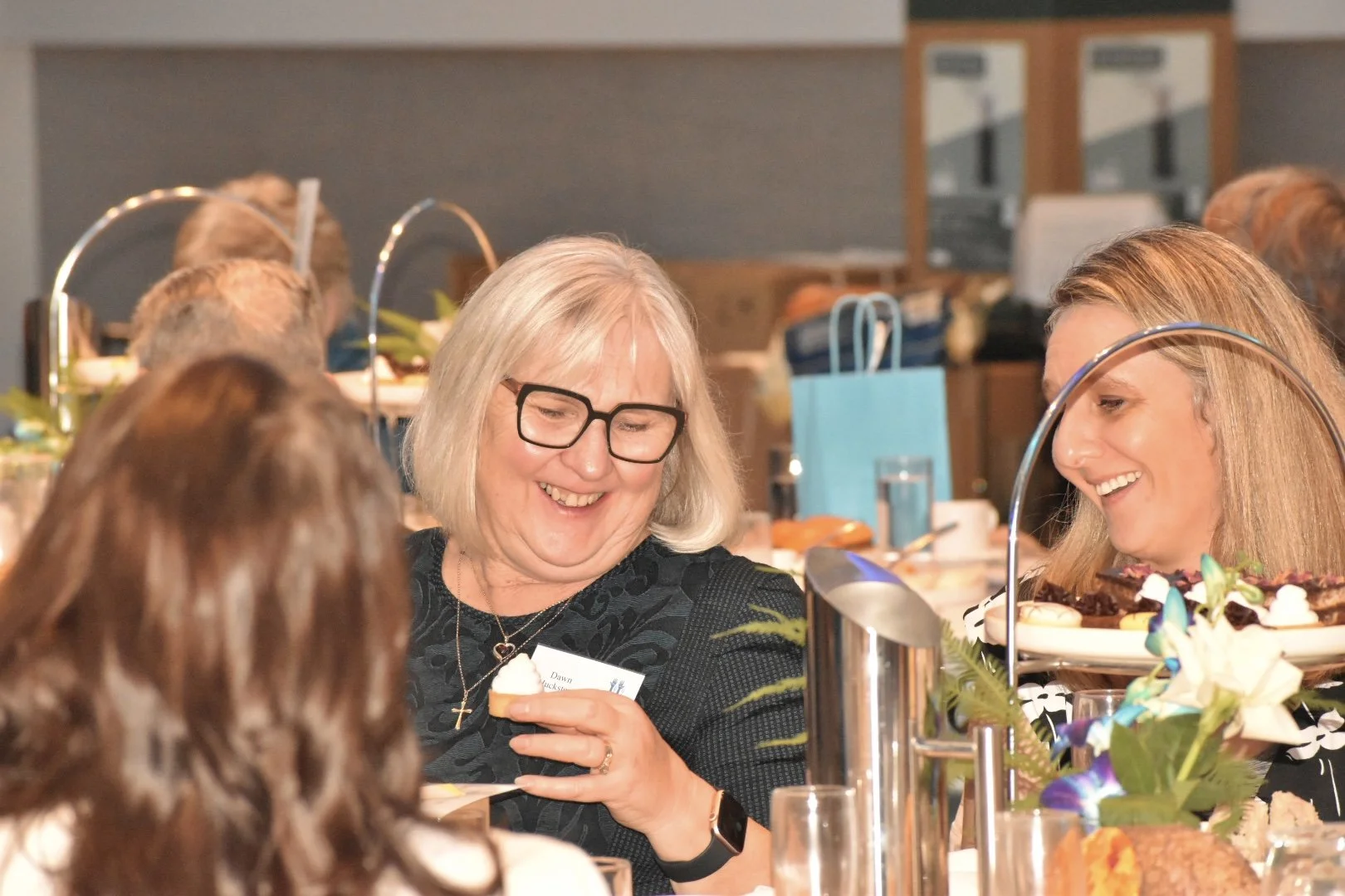 Group of women at a social gathering, smiling and chatting in a decorated indoor setting with food and flowers on the table.