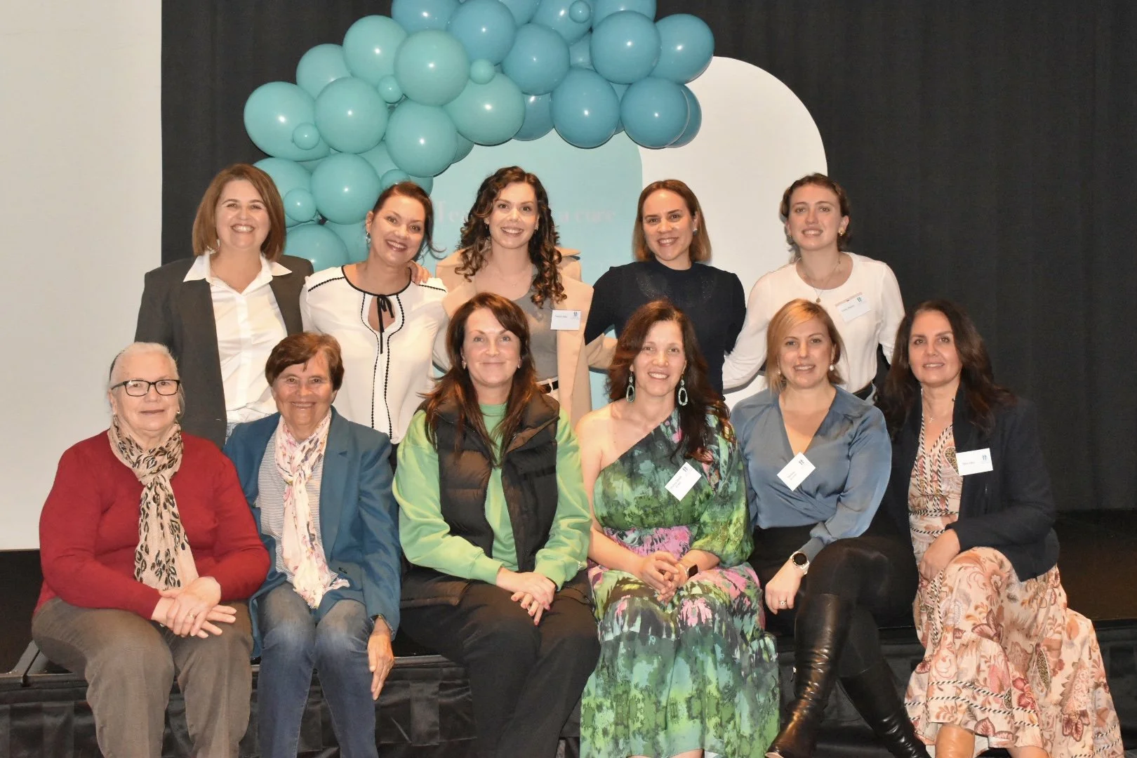 A group of 12 women posing on a stage in front of a decorative balloon backdrop, some standing and some seated, at a conference or event.