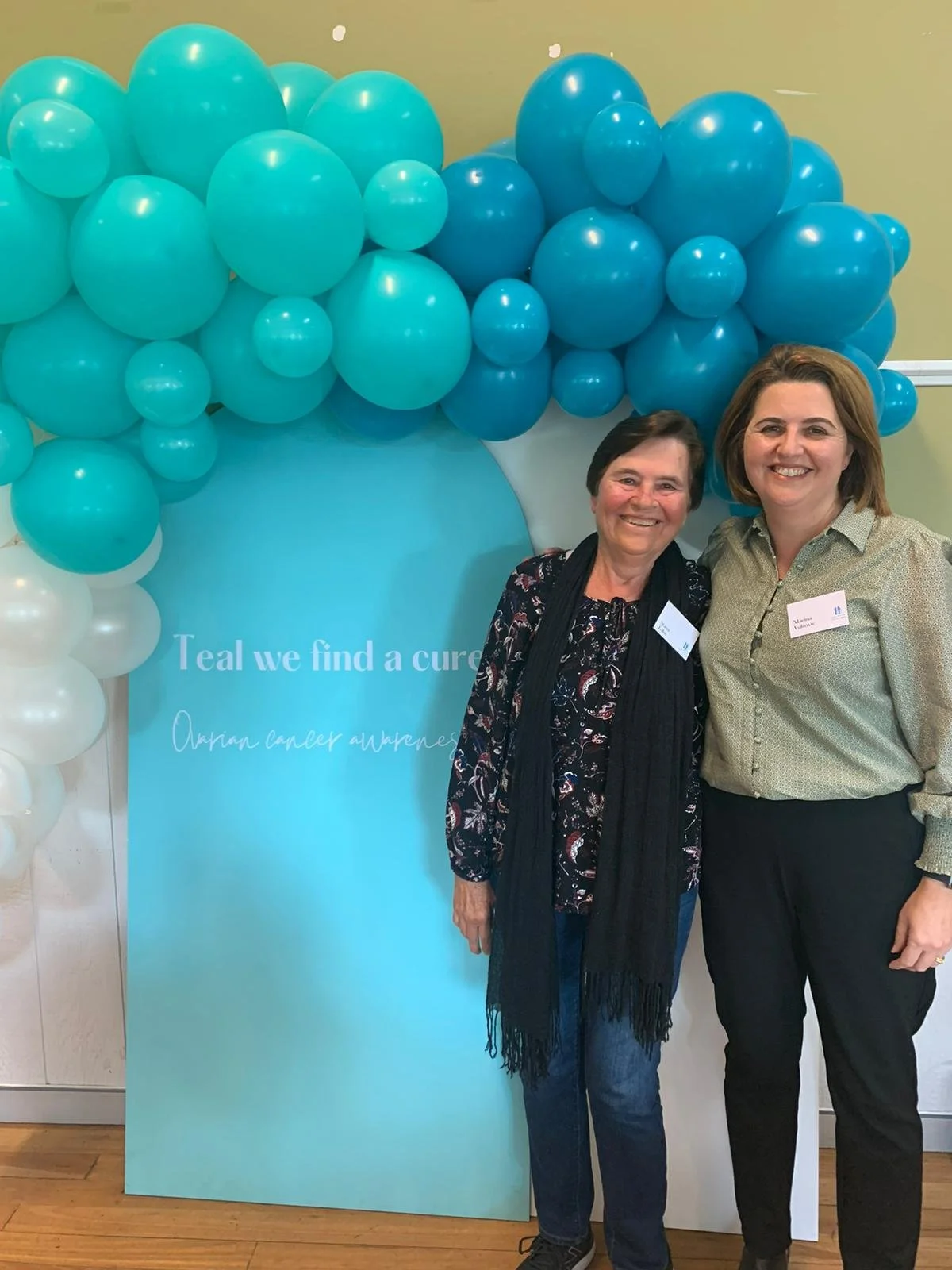 Two women standing together at a cancer awareness event with blue balloons and a blue sign that reads, 'Teal we find a cure. Ovarian cancer awareness.'