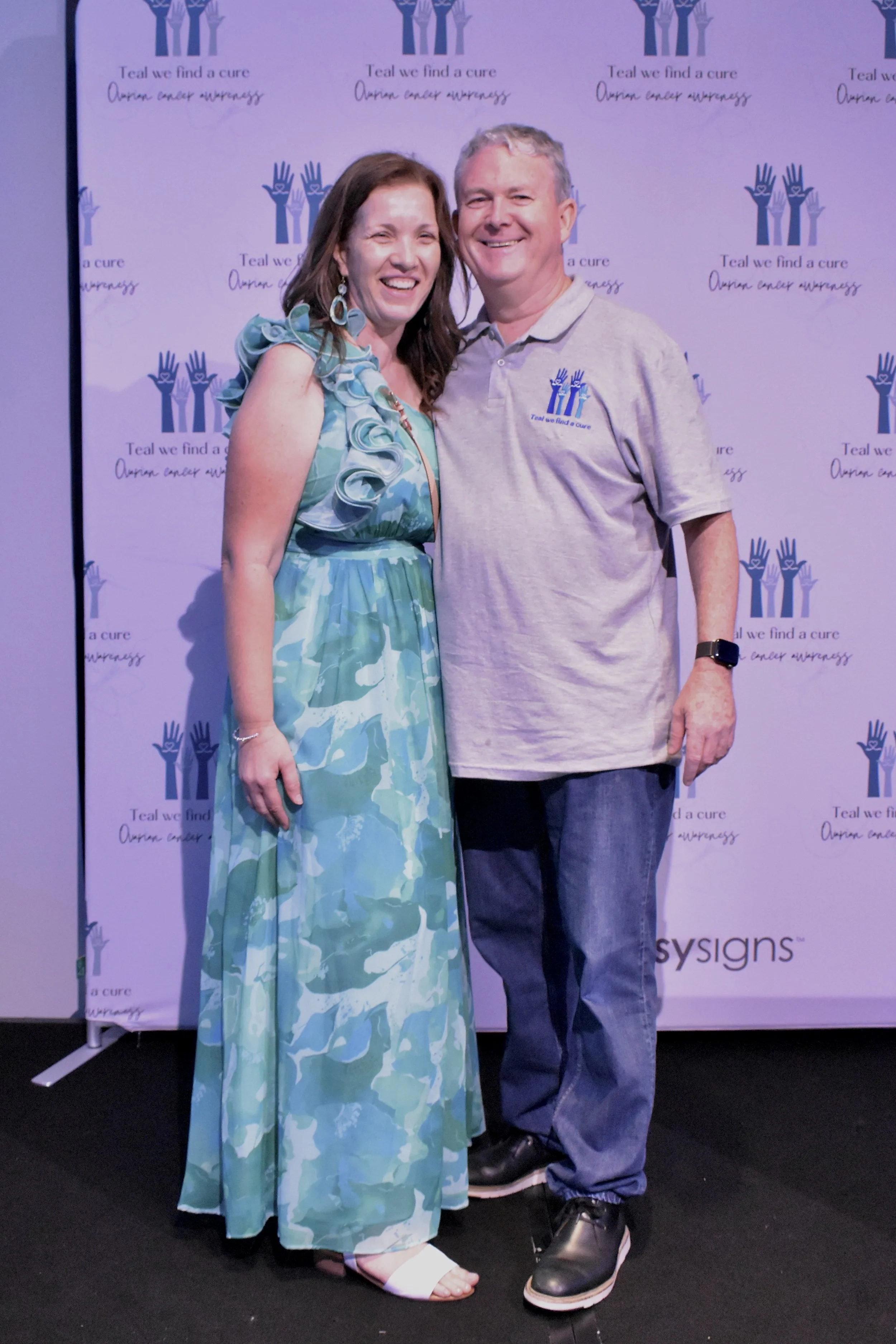 A woman and a man standing close together, smiling, in front of a backdrop with the text 'Teal we find a cure' and the phrase 'Oregon cancer awareness'. The woman is wearing a colorful, patterned maxi dress with ruffles on the shoulder, and the man i