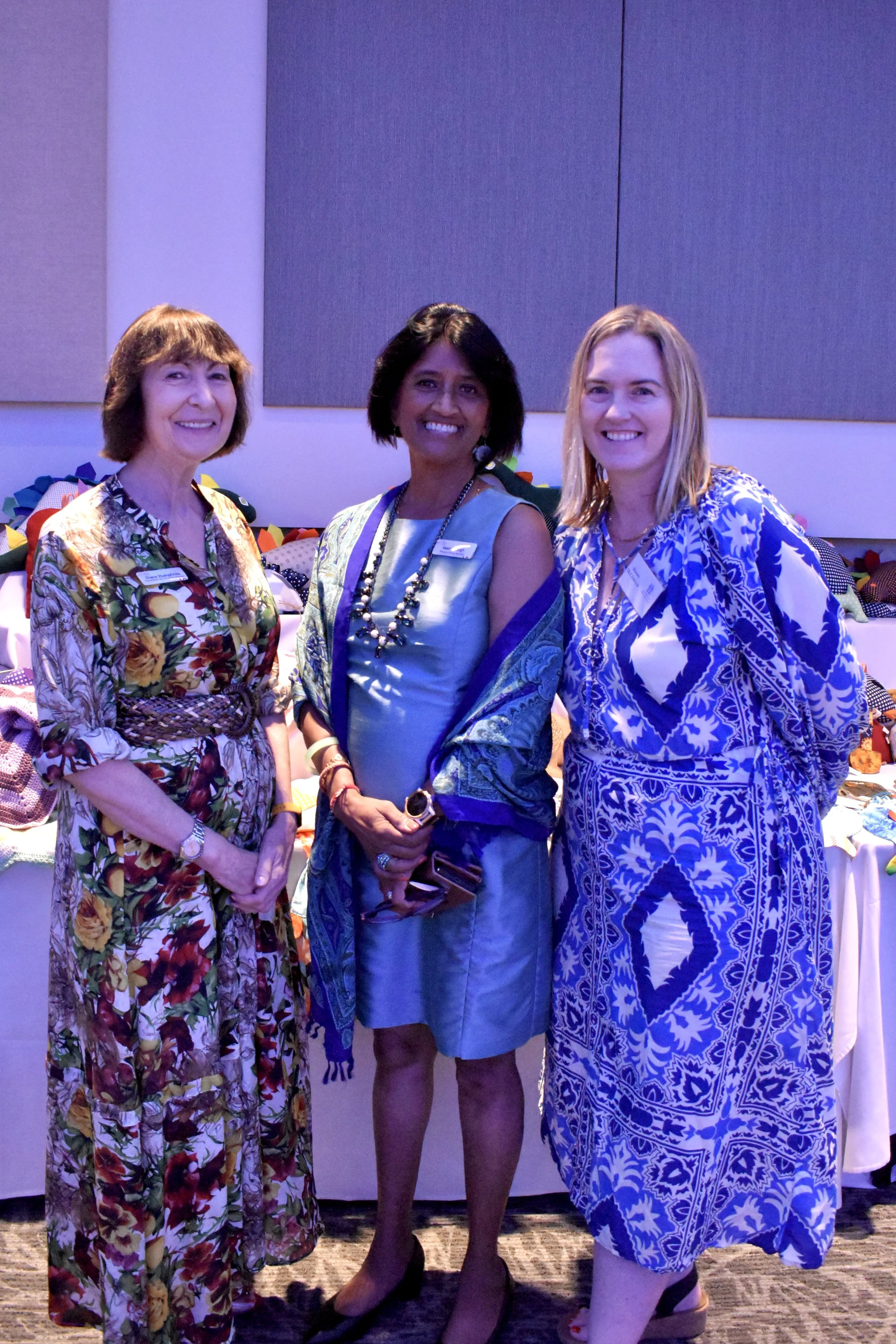 Three women standing together indoors at a social event, smiling and dressed in colorful, patterned dresses, with a table of fabric or craft items behind them.