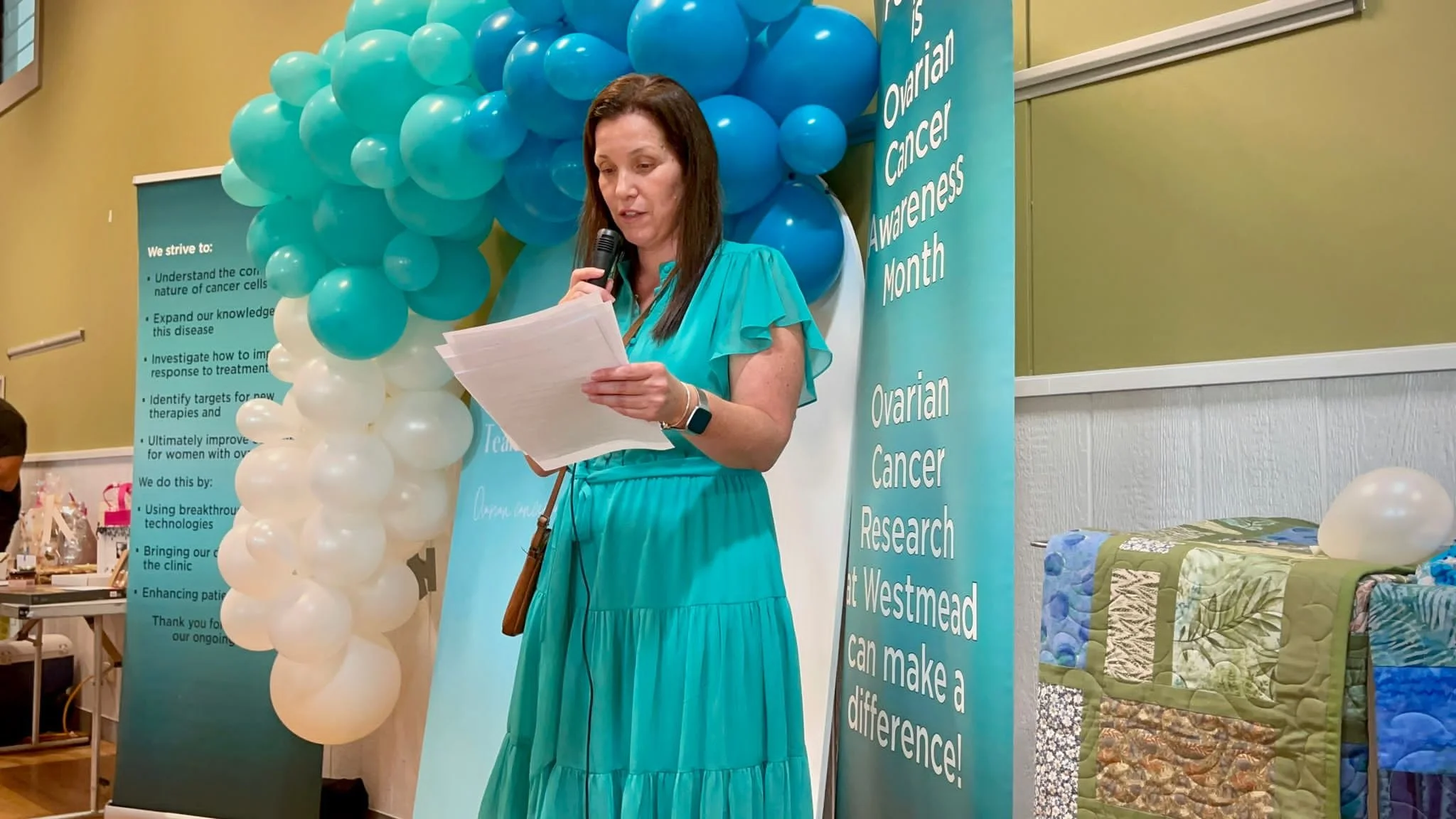 A woman in a teal dress speaking into a microphone and reading notes at a breast cancer awareness event, with teal and white balloons behind her and informational banners for ovarian and breast cancer research.