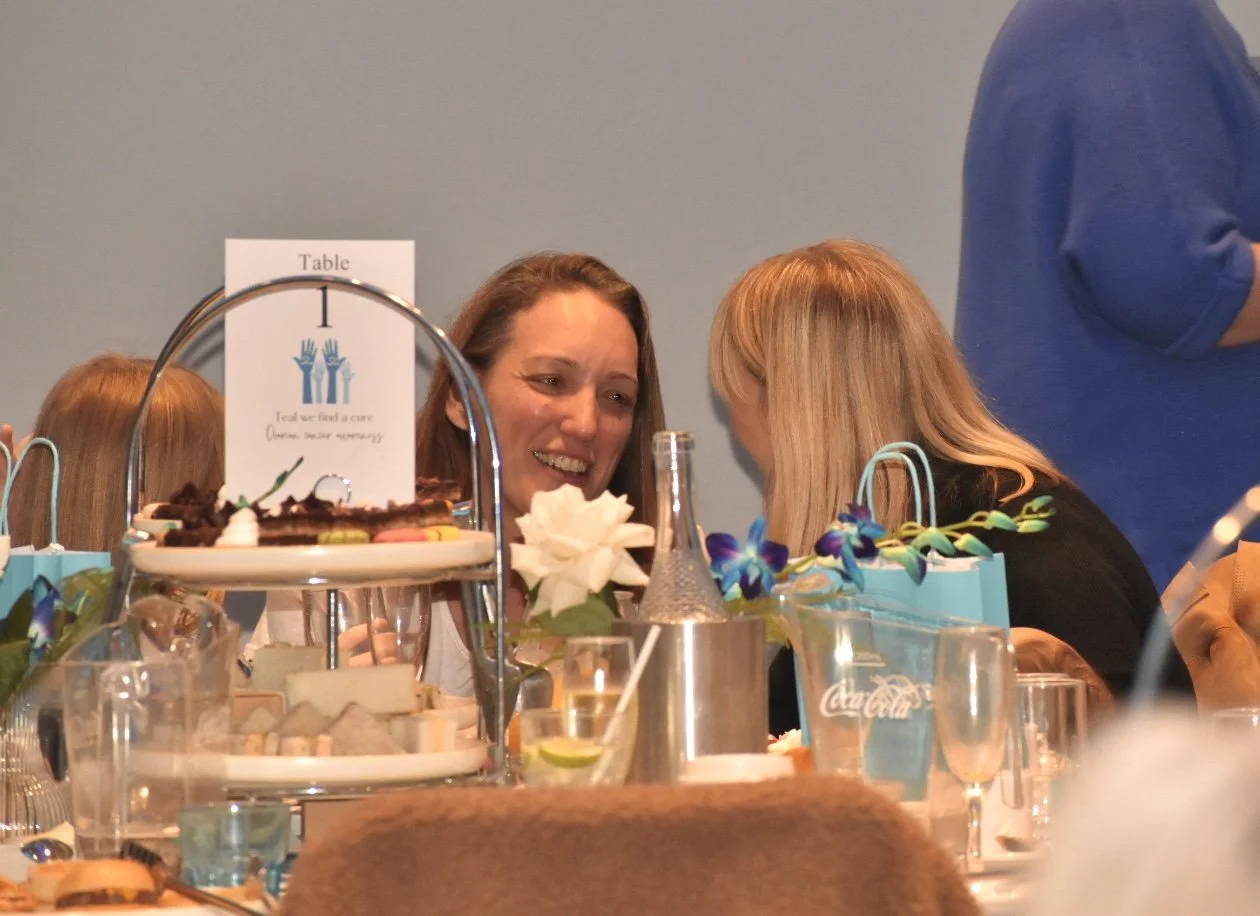 Two women seated at a table, smiling and engaged in conversation. The table is decorated with flowers, desserts, and beverages, with a sign in the background that says "Table" and has a graphic of two raised hands.