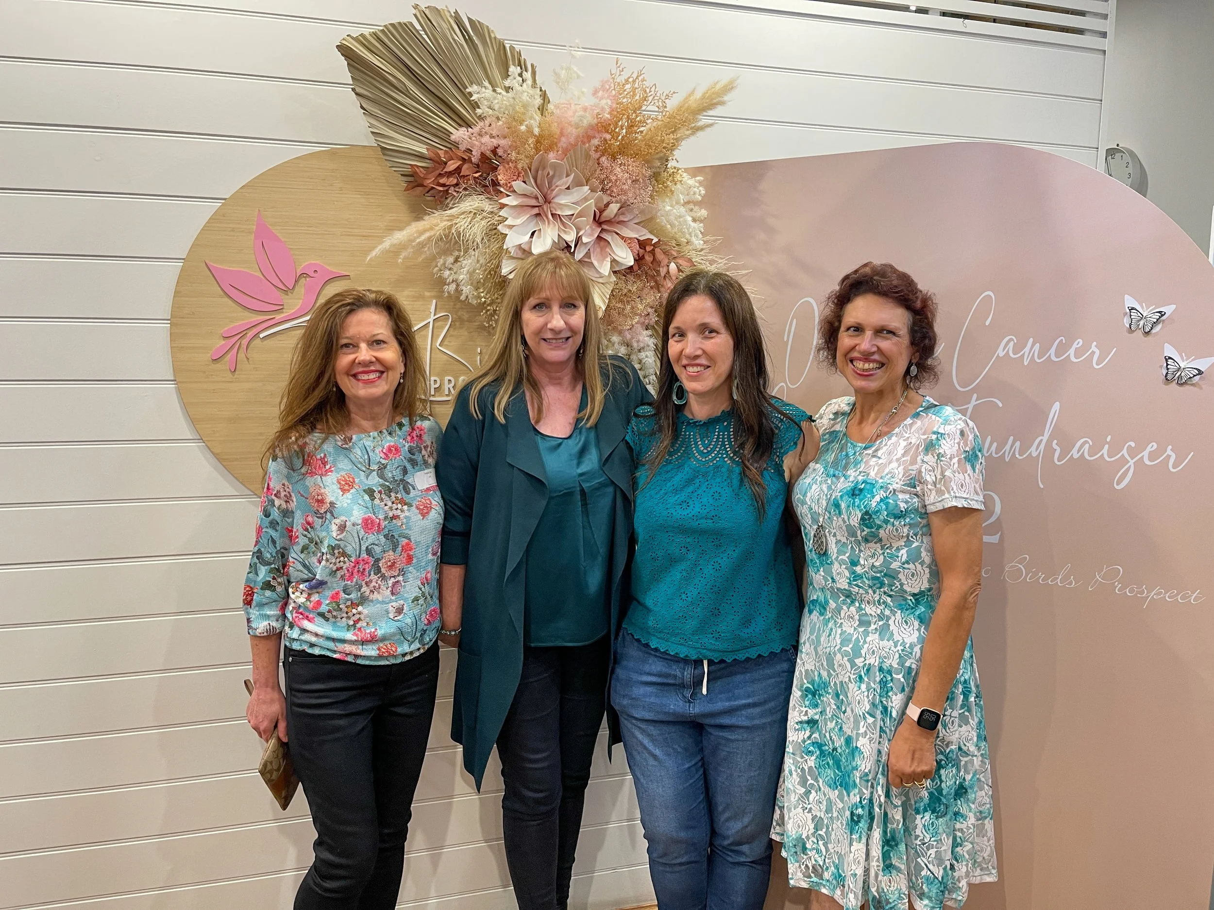 Four women standing together at a breast cancer fundraiser event, smiling in front of a decorative backdrop with pink and white flowers, a logo with a pink bird, and text about the event.