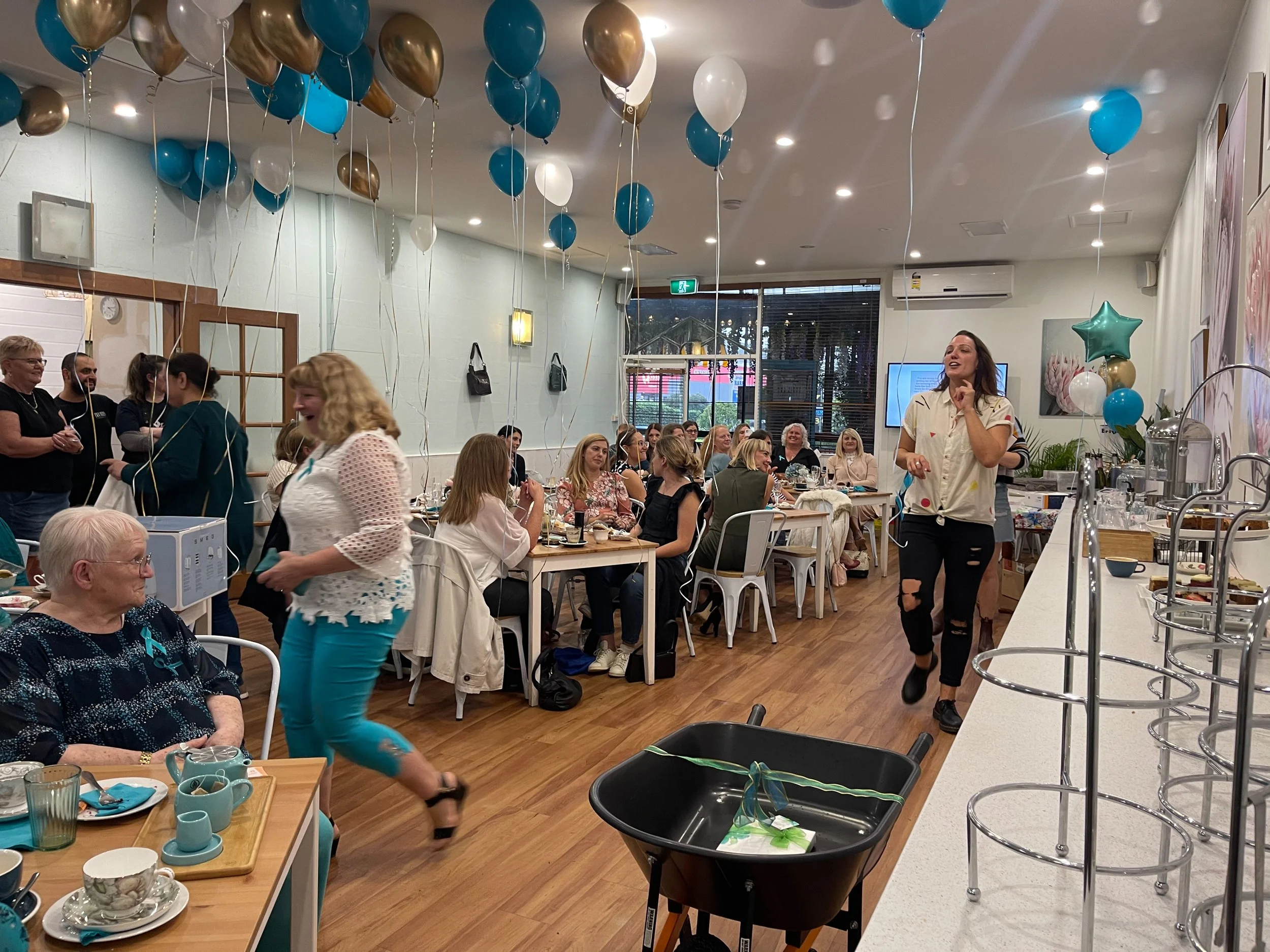 A gathering of people in a decorated indoor space with blue, white, gold, and star-shaped balloons, some of which are floating and tied to the ceiling. Attendees are seated at tables or standing, enjoying food and conversation. A woman in a white jac