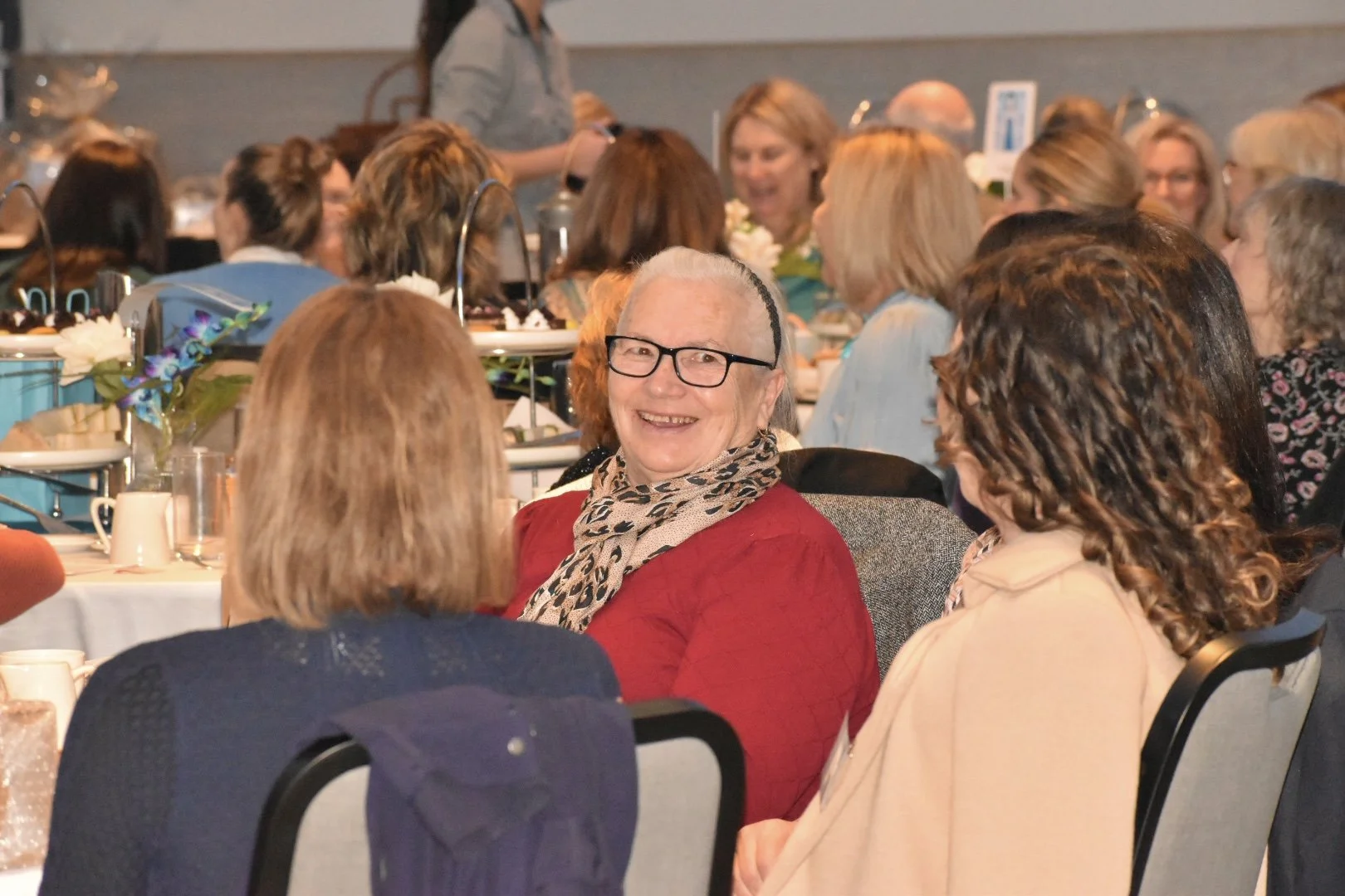 A group of women at a social event, with one elderly woman in the center smiling and wearing glasses, a red top, and a leopard print scarf, sitting at a dining table with floral decorations, in a large room filled with many other women.