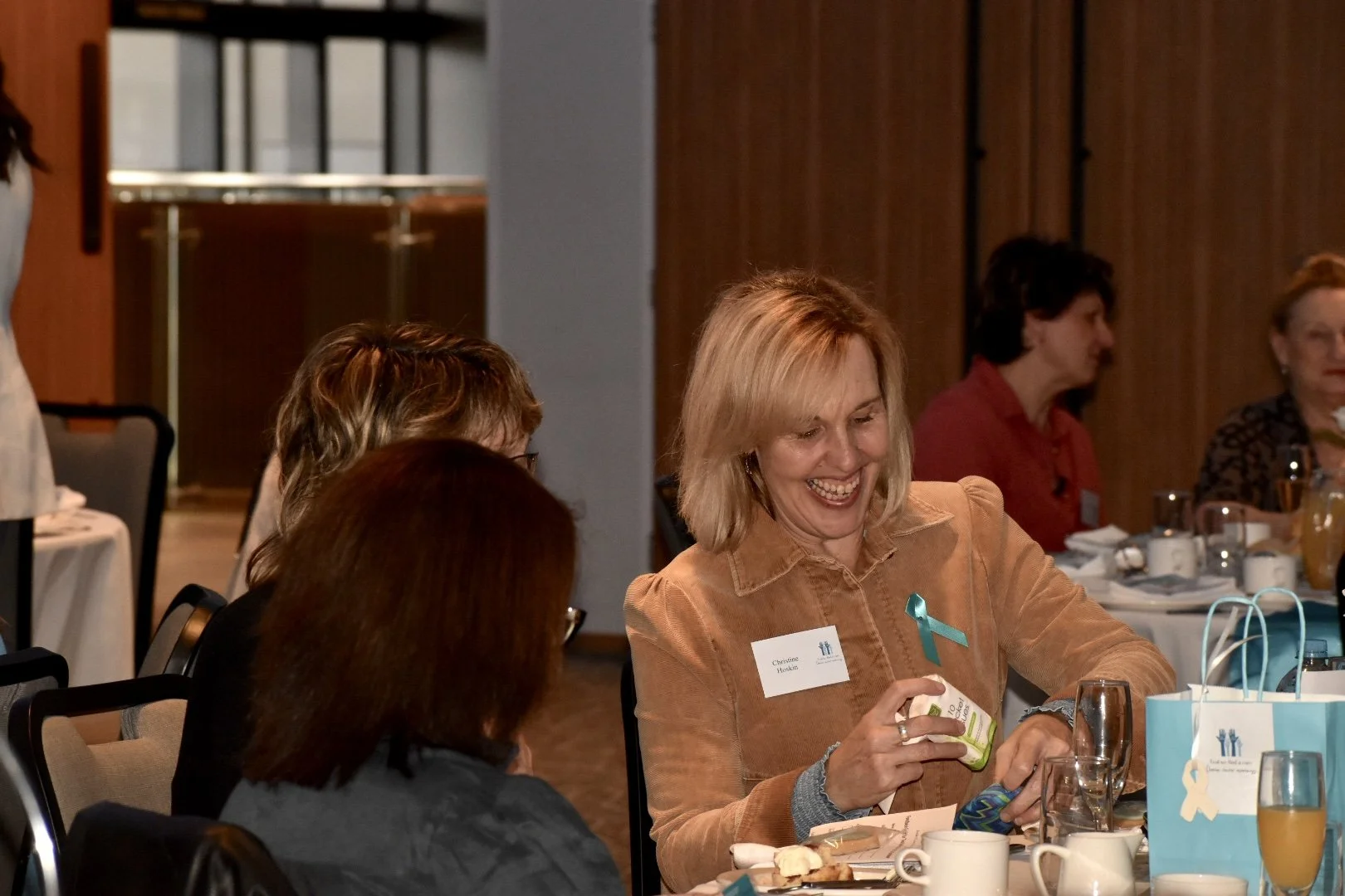 Women sitting at a round table during a formal event, one woman with blonde hair smiling and holding a gift, with gift bags and drinks on the table.