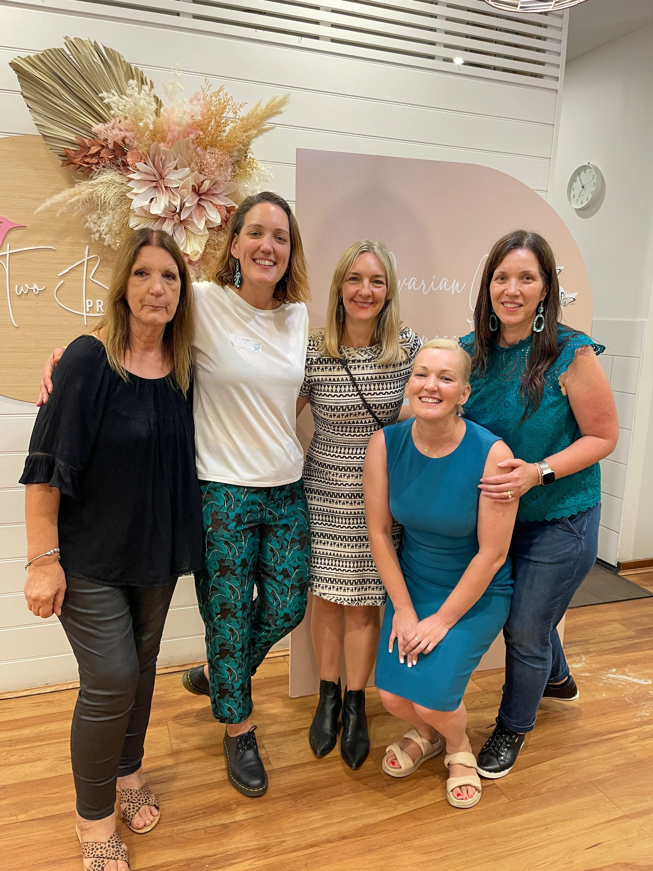 Five women standing together indoors, smiling for a group photo with decorative pink and beige flowers and a beige background featuring cursive writing.