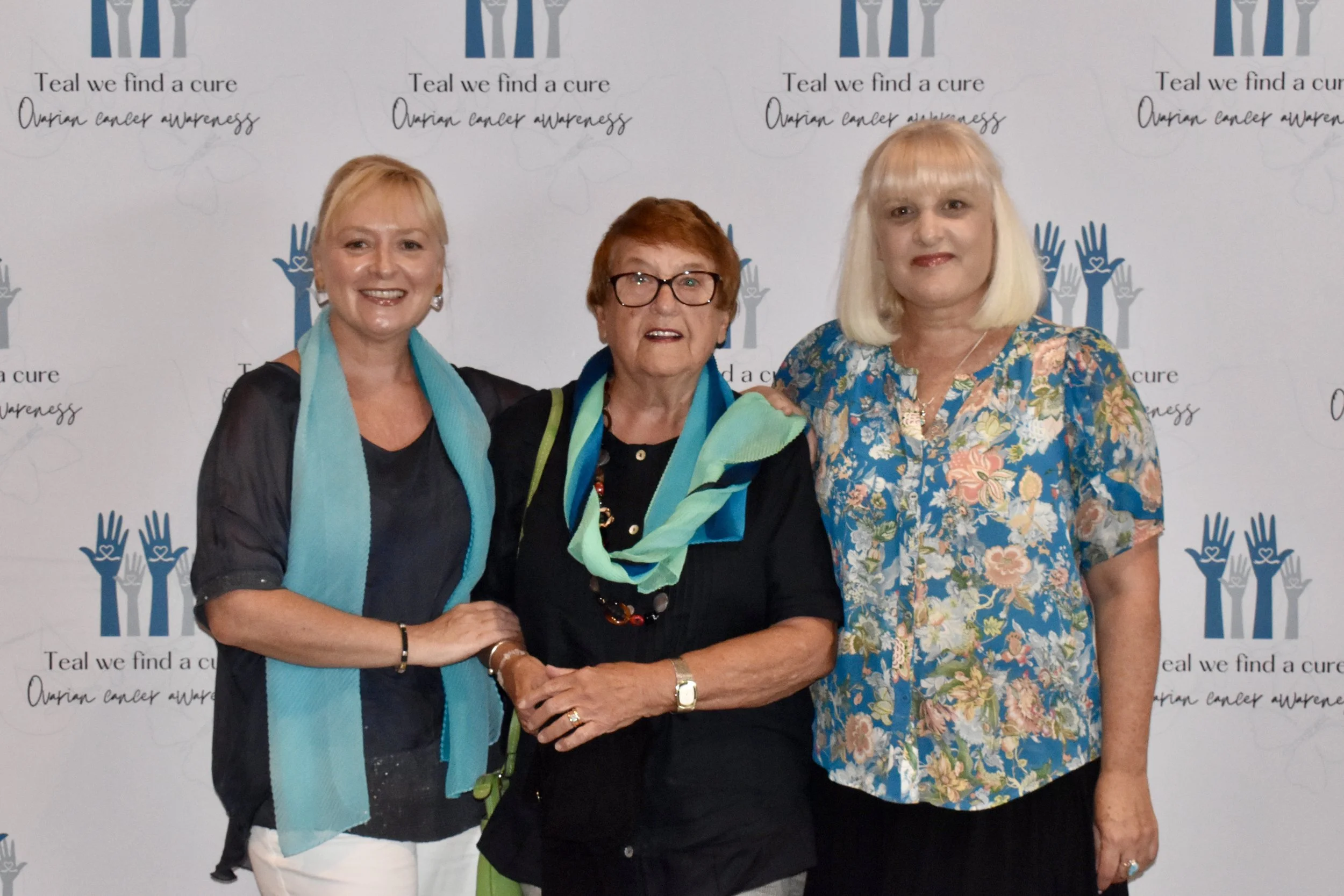 Three women standing together at a breast cancer awareness event in front of a backdrop with the text 'Teal we find a cure' and symbols of teal and gray hands. The women are smiling and dressed in casual and floral clothing.