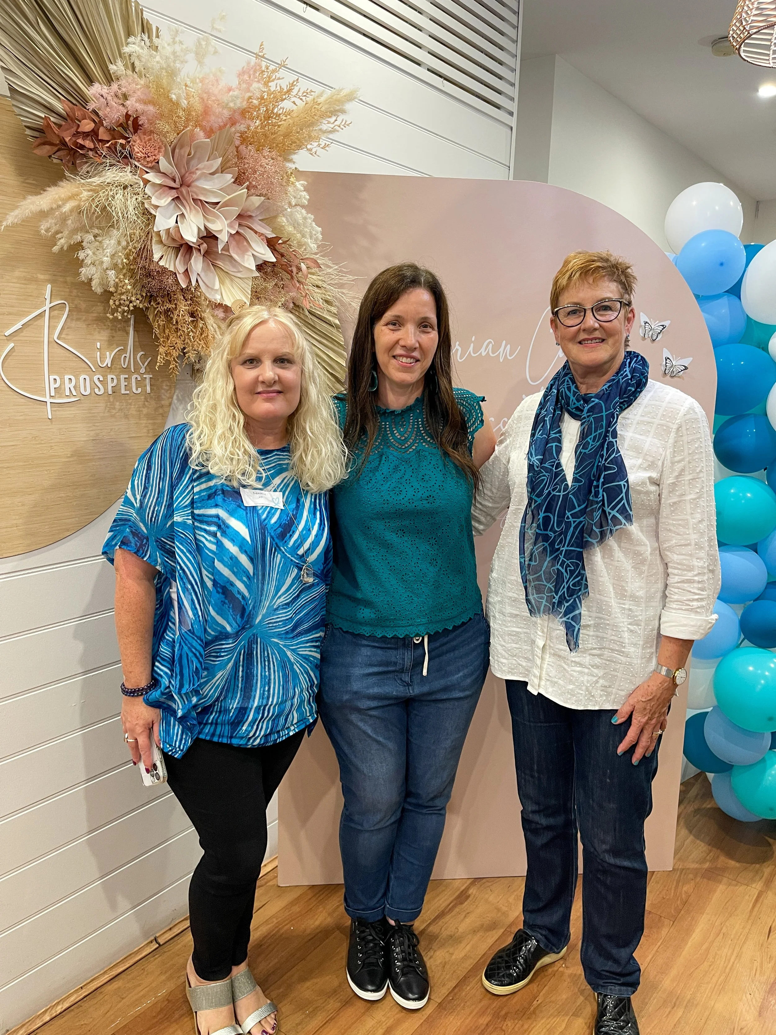 Three women standing together in front of a decorative backdrop with balloons, flowers, and butterfly accents, smiling at the camera.