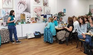 A woman presents to a group of women seated at tables in a gallery or classroom setting, with artwork on the walls.