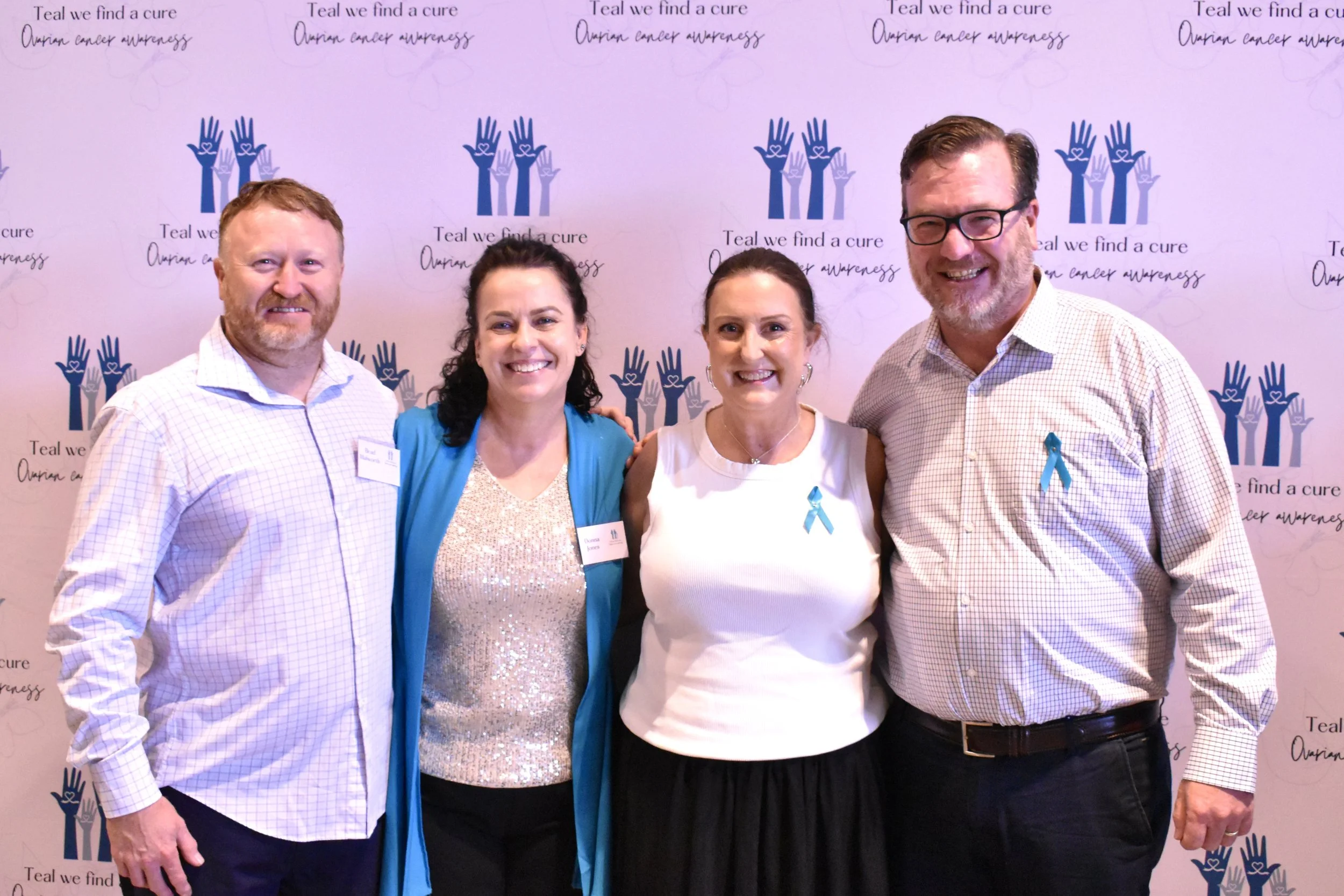 Four people standing together smiling at a fundraising event with a pink backdrop featuring blue and purple hand designs and teal ribbons, supporting ovarian cancer awareness.