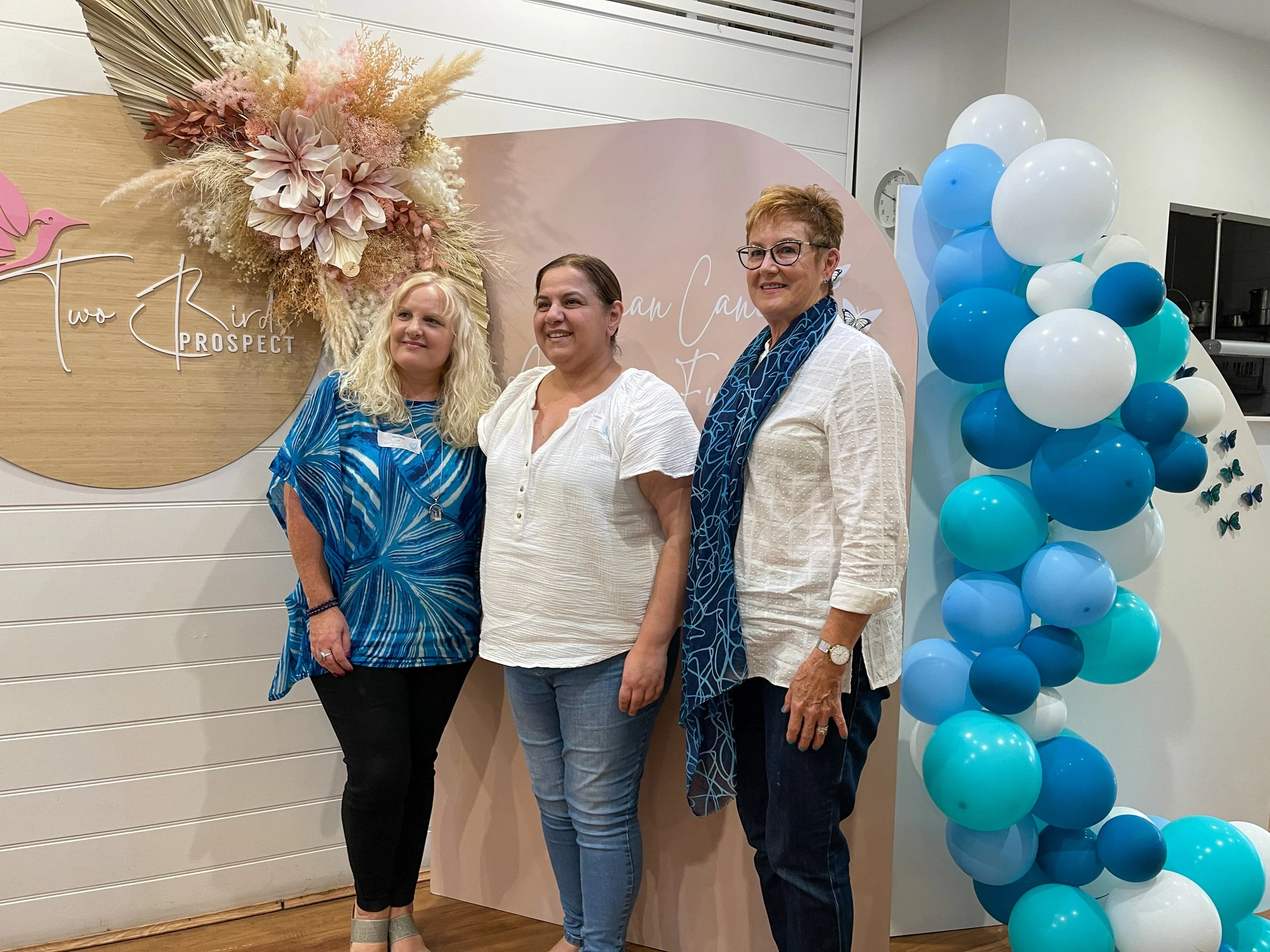 Three women standing together at a celebration event, with a decorative backdrop and balloons. The backdrop has a floral arrangement and text that reads 'Two Pink Prospects'. The women are smiling and dressed casually, with one wearing glasses and a 