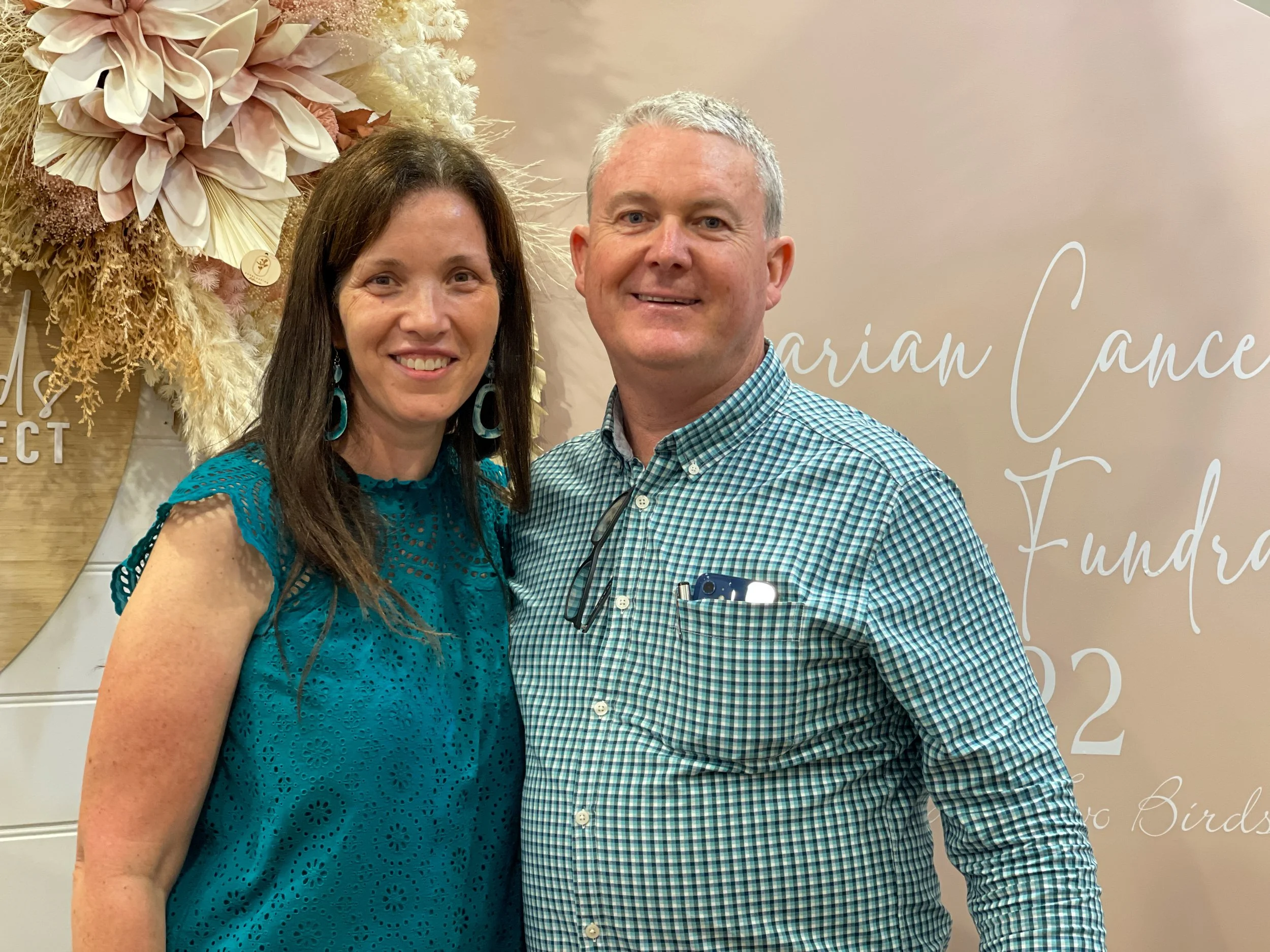 A smiling woman and man posing together in front of a beige backdrop with white cursive text. The woman is wearing a teal eyelet dress and earrings, and the man is wearing a green and blue checkered shirt with glasses hanging from his collar, and has