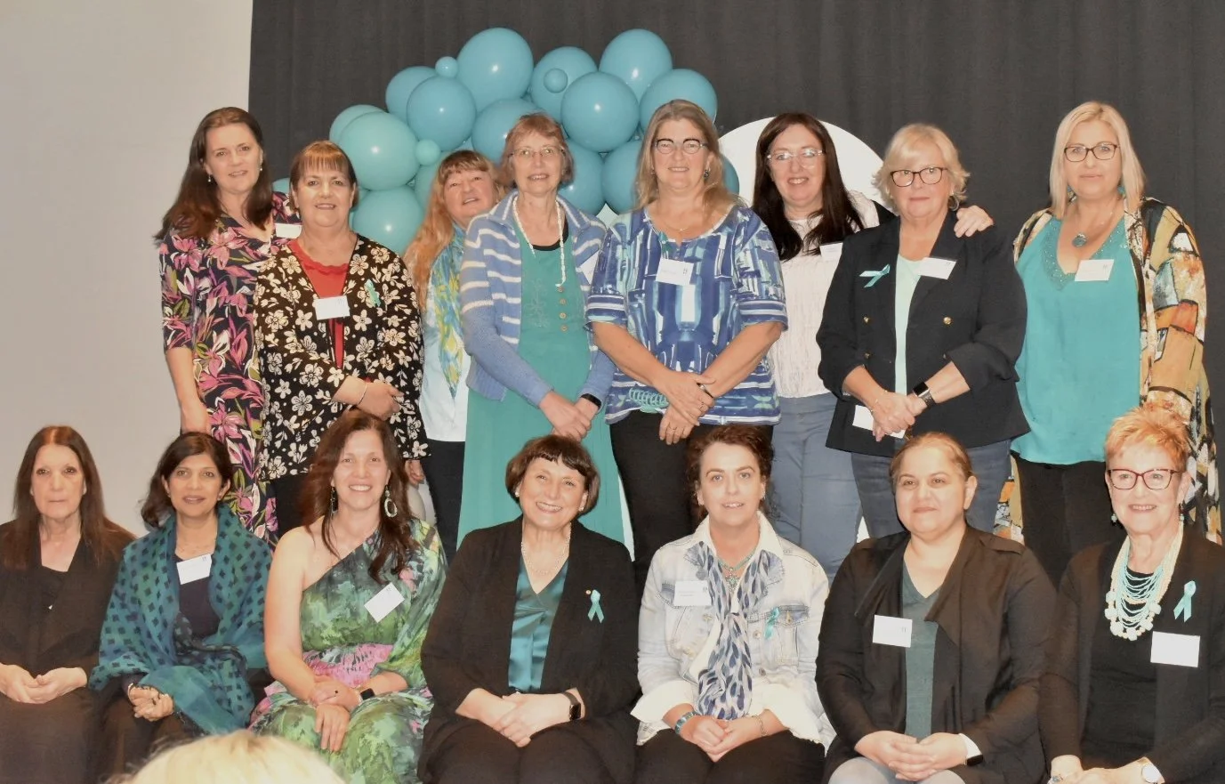 Group of women at a conference or event, standing and sitting in front of a backdrop with teal balloons.