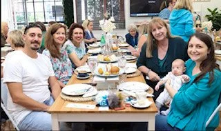 Group of people sitting at a long dining table, smiling, with children and adults, in a bright indoor setting.