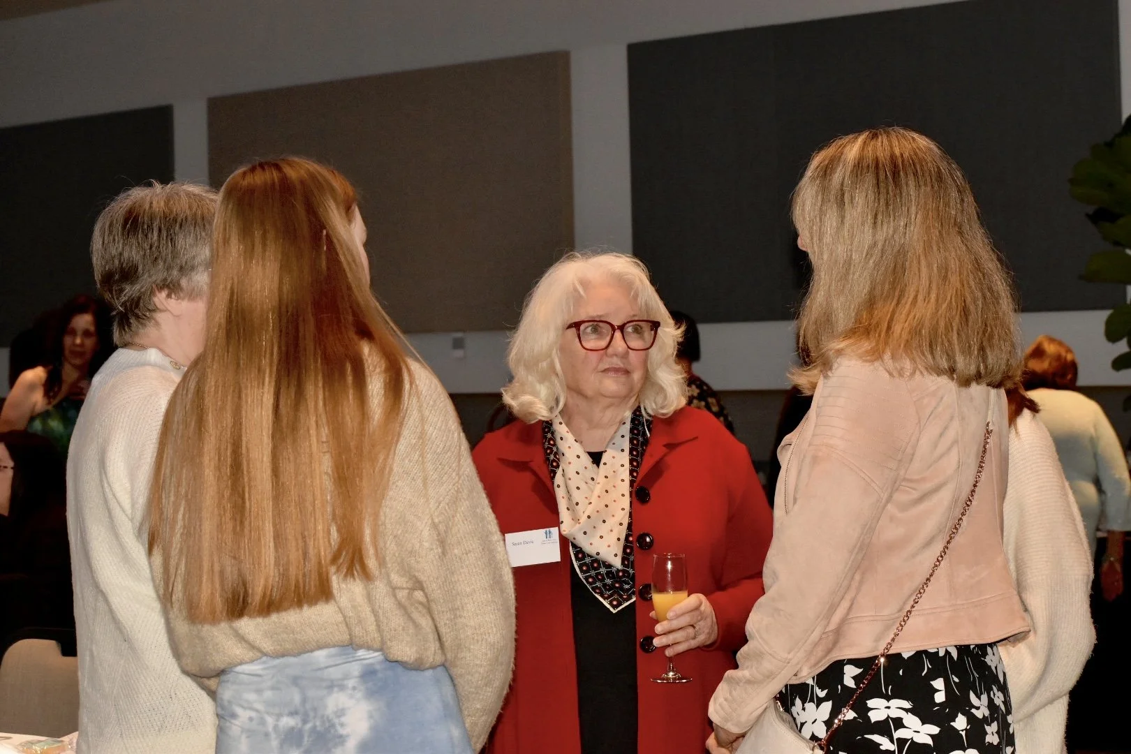 Four women are standing and talking at a social gathering. One woman in a red blazer with white hair and glasses is holding a glass of champagne. The other women have different hair colors and styles and are facing her.