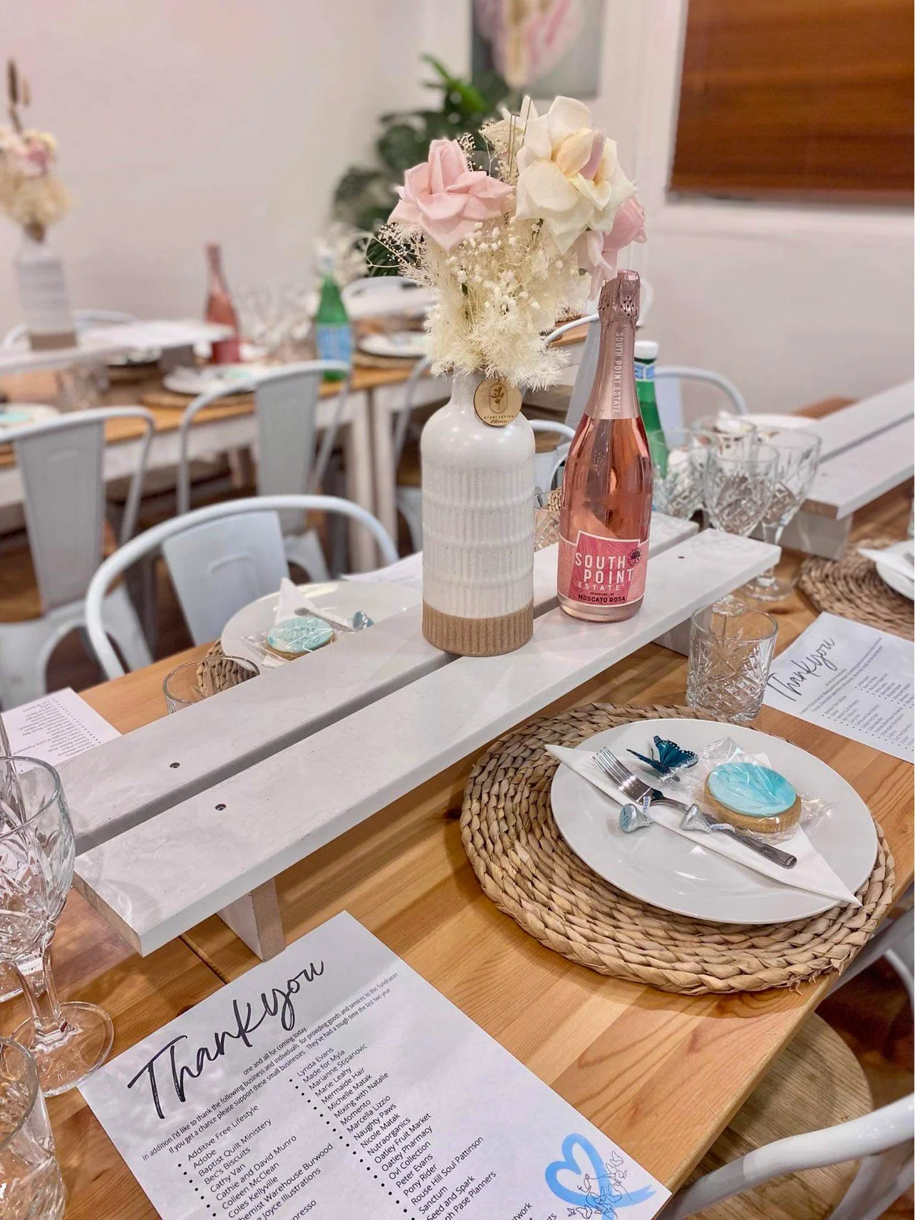 A table decorated for an event with a white basket vase holding pink and white flowers, a pink sparkling rosé bottle, and place settings with woven placemats, white plates, clear glasses, and decorated cookies.