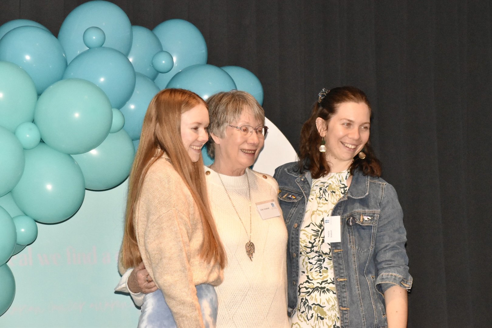 Three women smiling and standing together at an event, with a background of teal balloons and a dark backdrop.