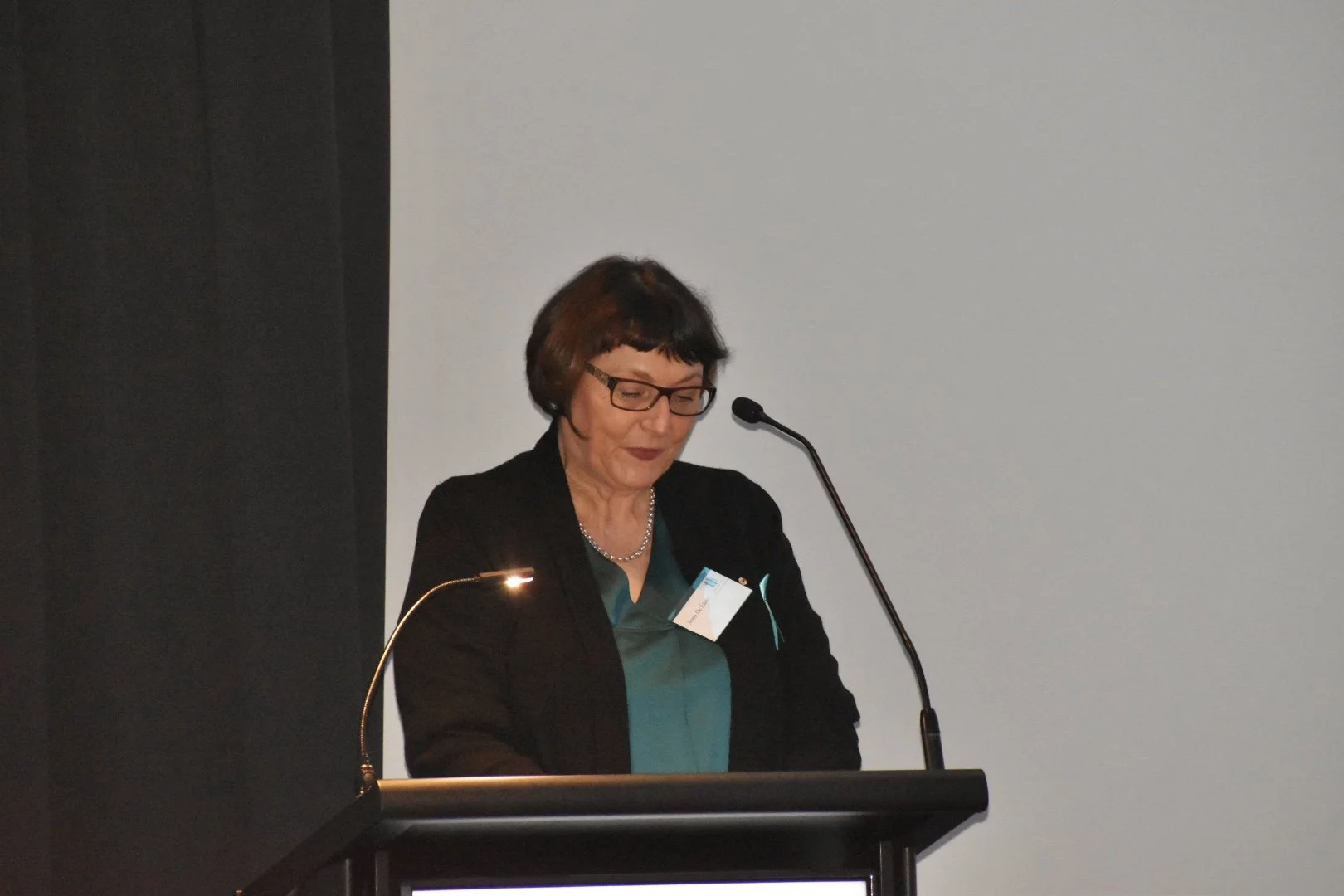 A woman with short dark hair, glasses, and wearing a dark blazer and green blouse, is standing at a podium with a microphone, looking down at notes during a speech or presentation.
