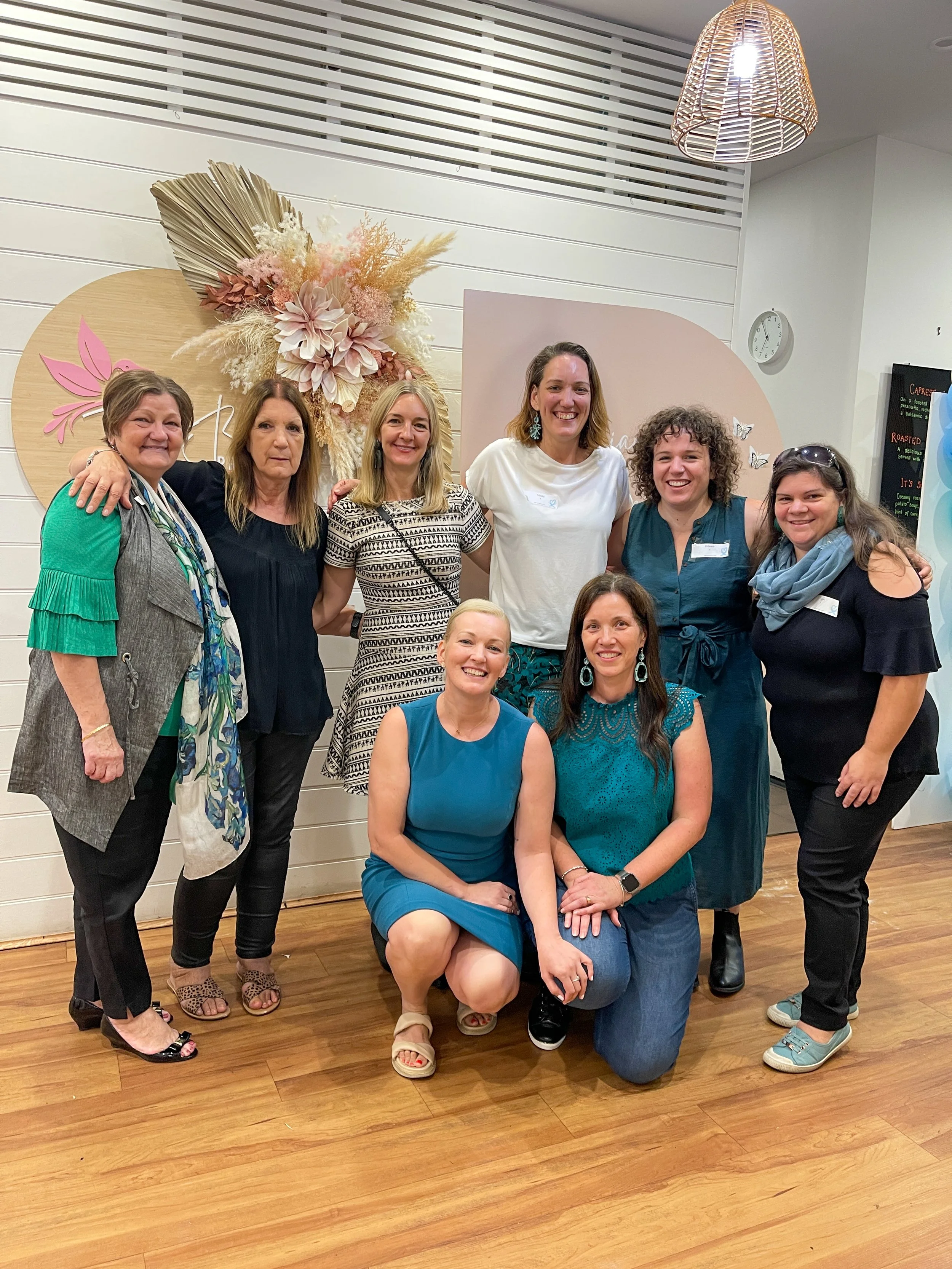 Group of nine women posing together indoors, smiling at the camera, with a decorated floral backdrop and a white wall with a clock in the background.