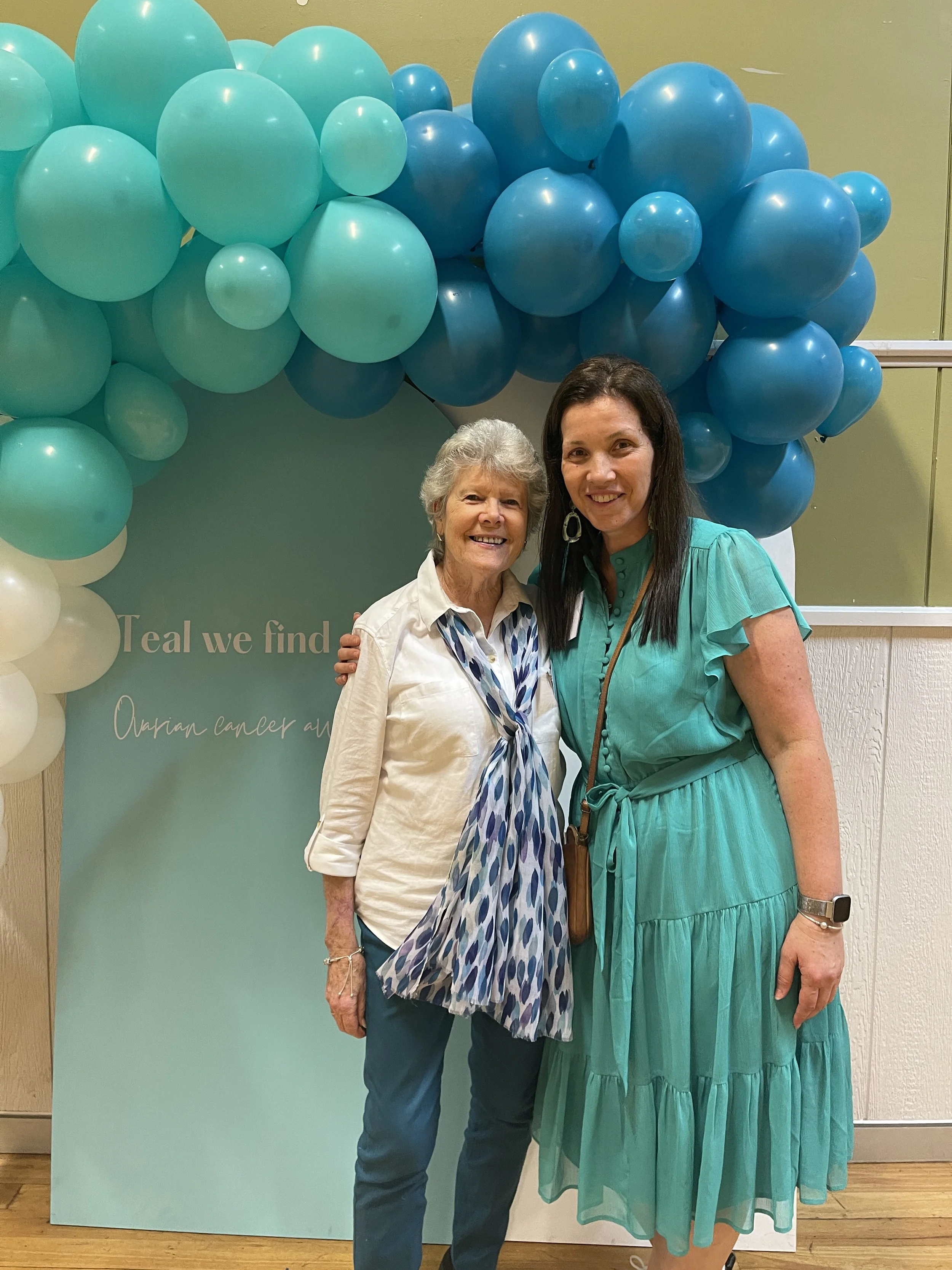 Two women standing together, smiling, in front of a teal and white balloon arch, with a teal sign that reads 'Teal we find' and 'Ovarian cancer awareness' in the background.